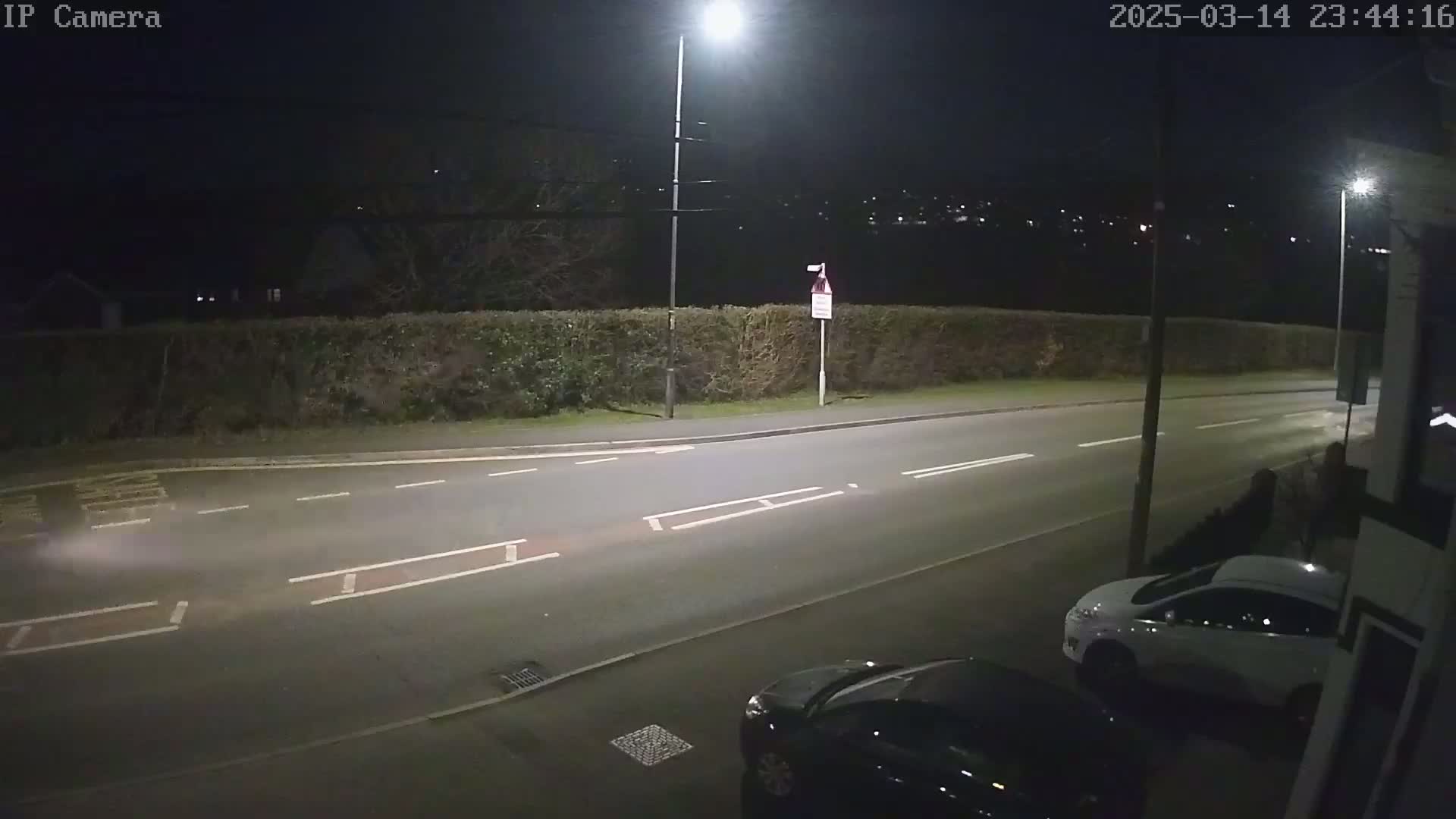 A nighttime view of a street with two parked cars, illuminated by streetlights, bordered by a hedge.