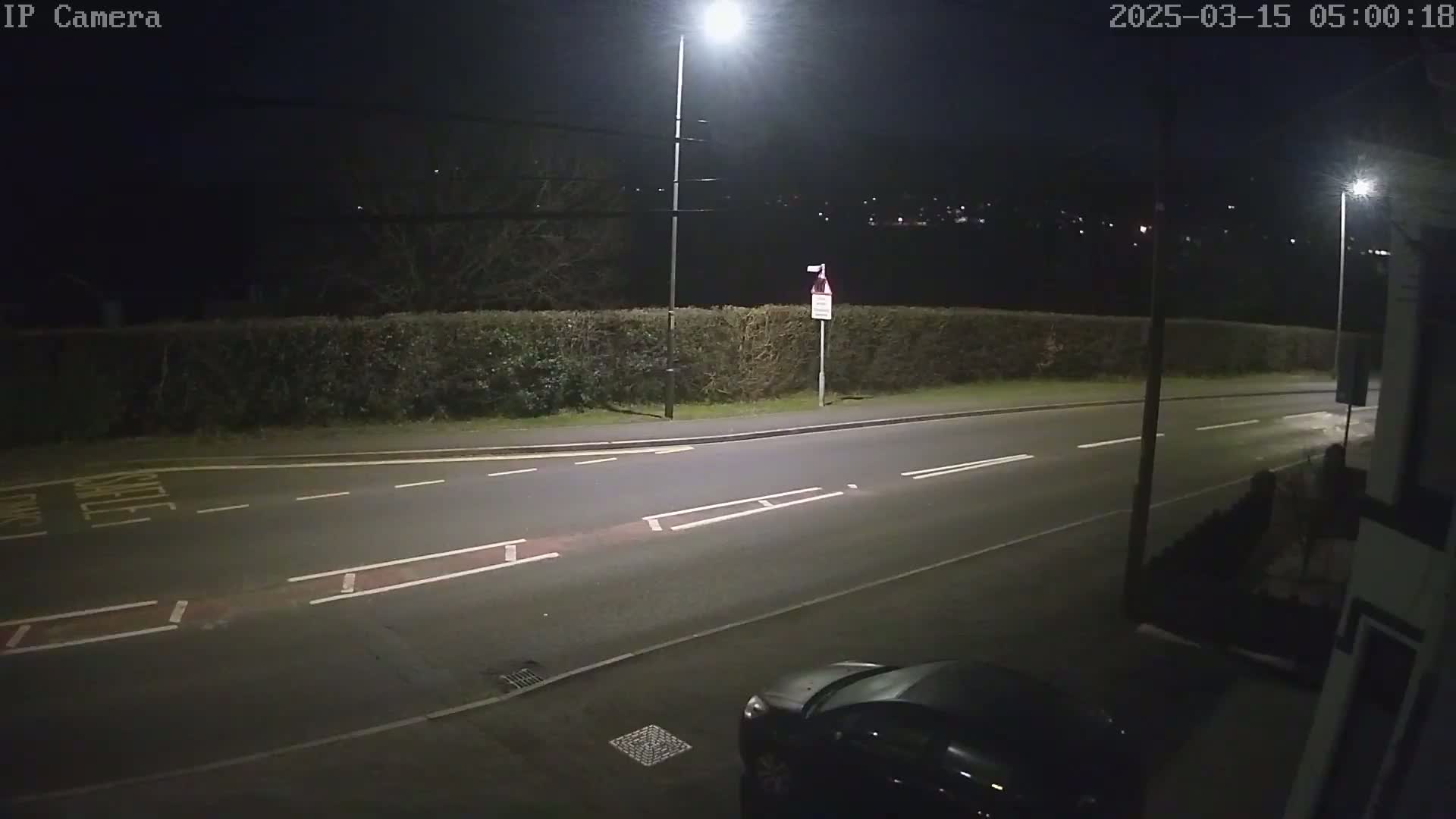 A dark, empty road at night, lit by streetlights, is bordered by a hedge, with a car partially visible in the lower right corner.