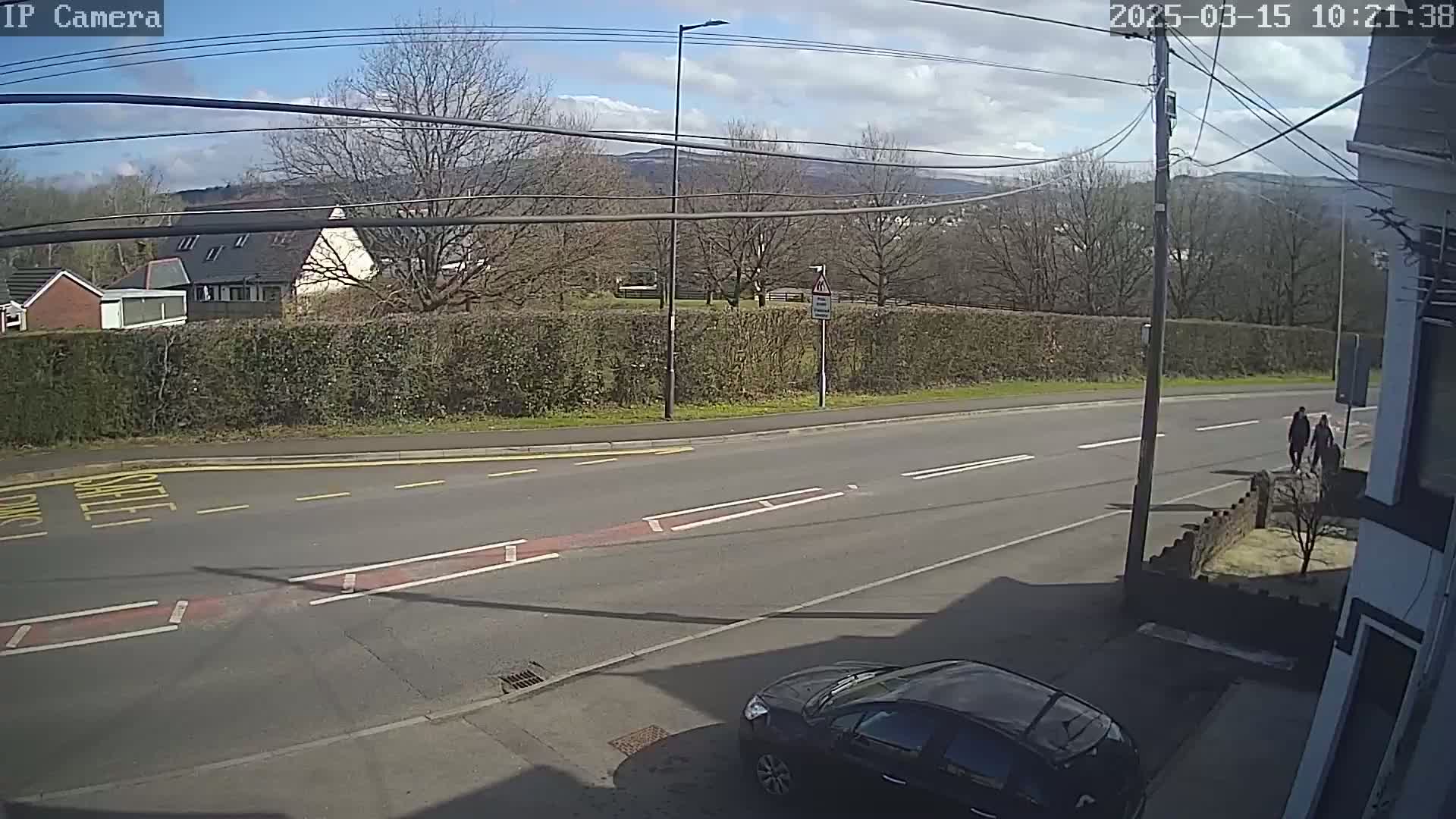 A sunny day reveals a residential street scene with a dark-colored car parked near a house, a hedge bordering the road, and pedestrians walking along a road that leads to hills in the distance.
