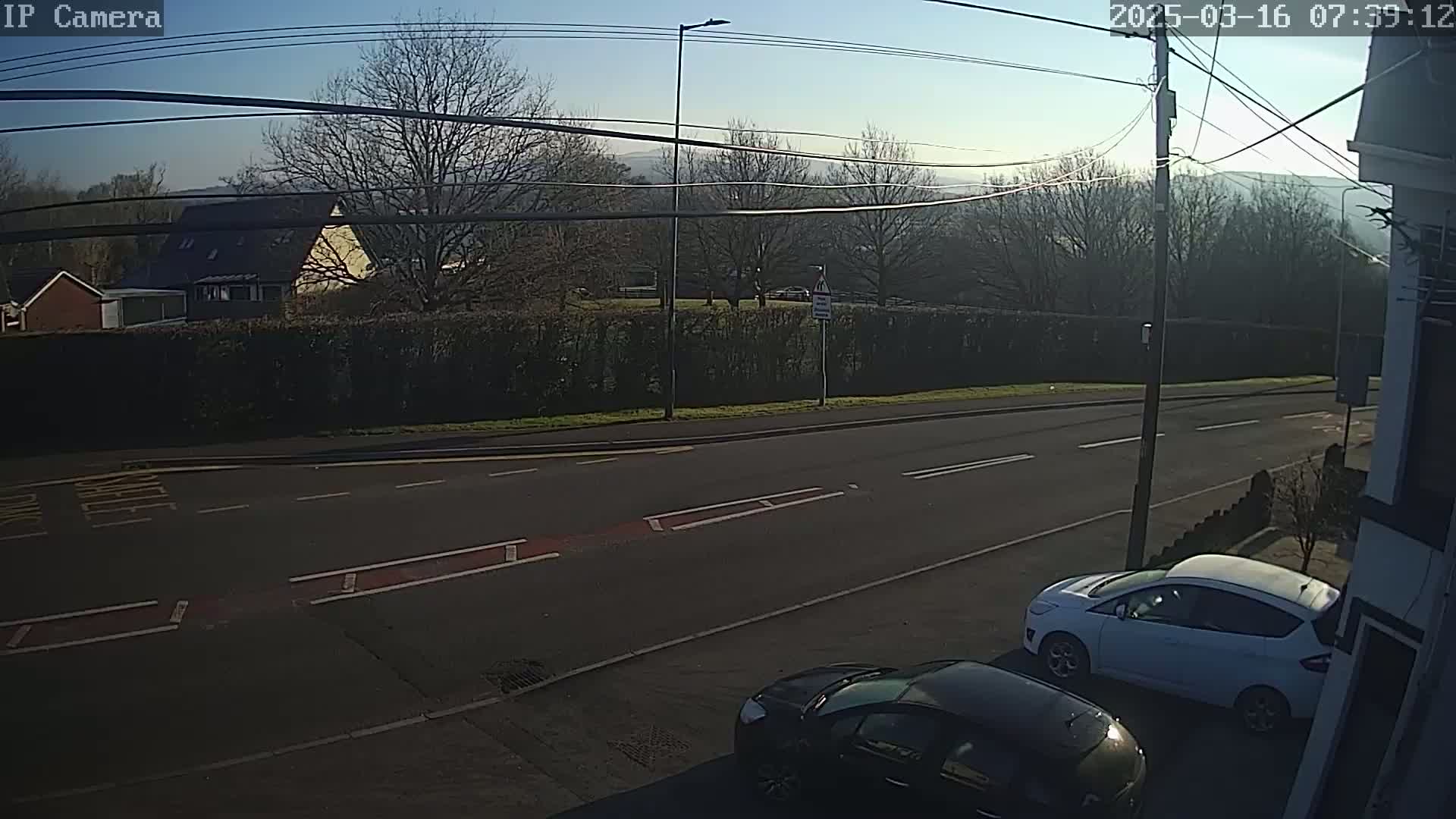 A sunny day reveals two cars parked on the side of a road, with a hedge and houses visible in the background.
