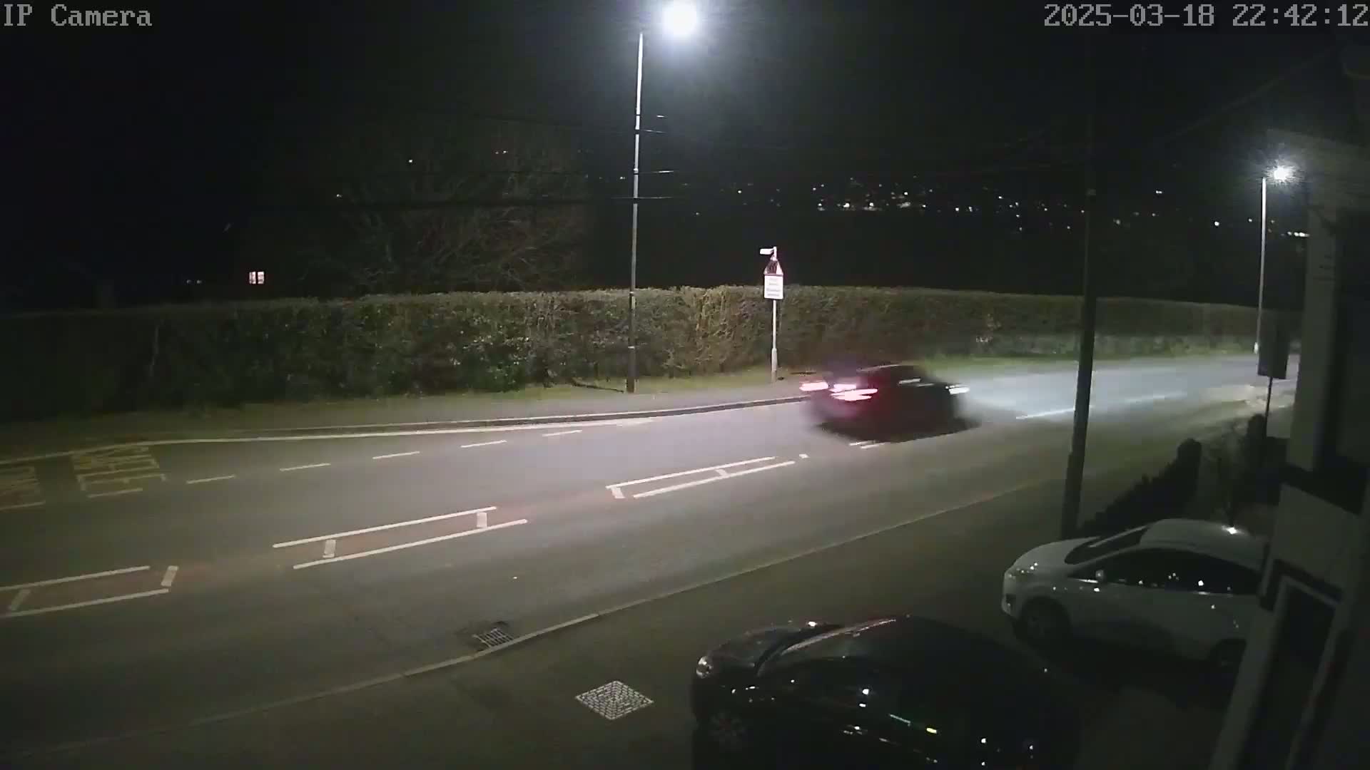 A dark-colored car drives on a road at night, past a parked white car, under illuminated streetlights.