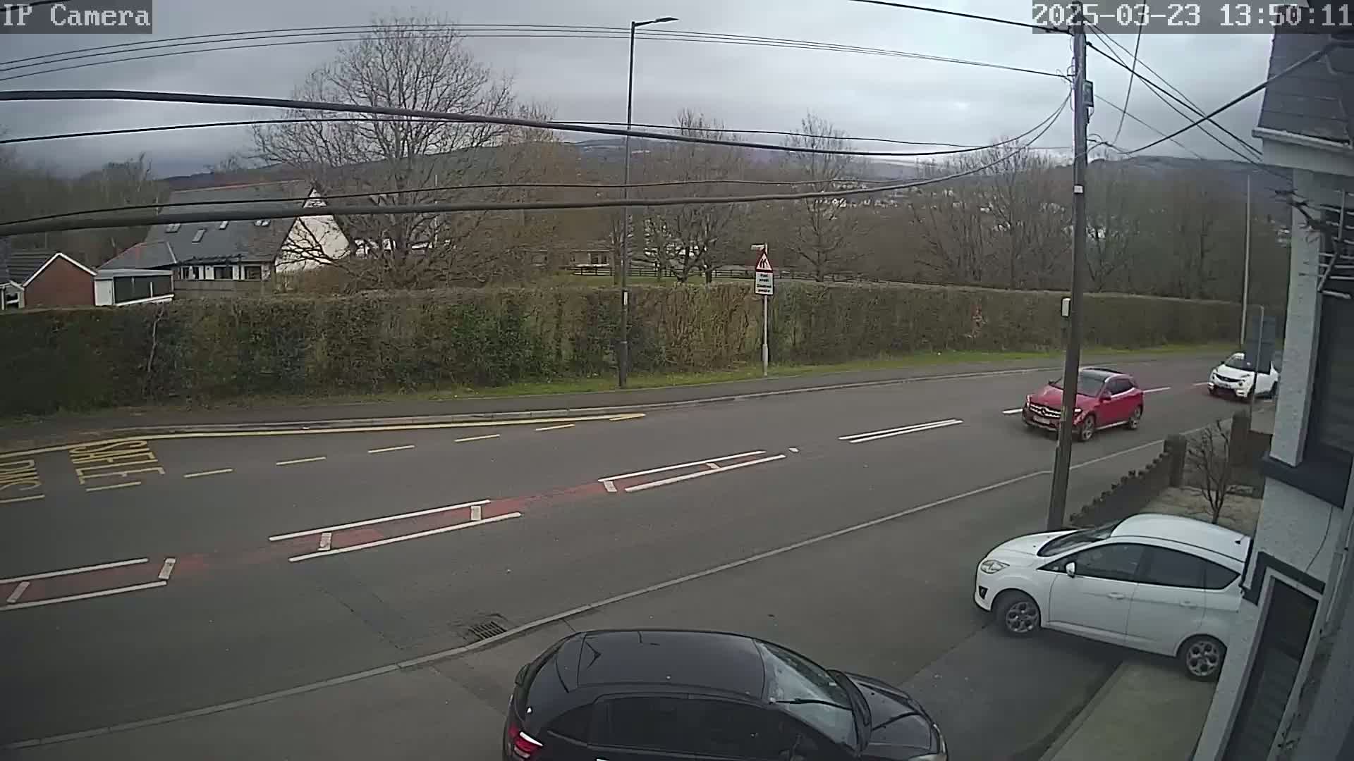 A street scene on an overcast day shows a road with several cars parked and driving, bordered by a hedge and houses, with hills visible in the distance.