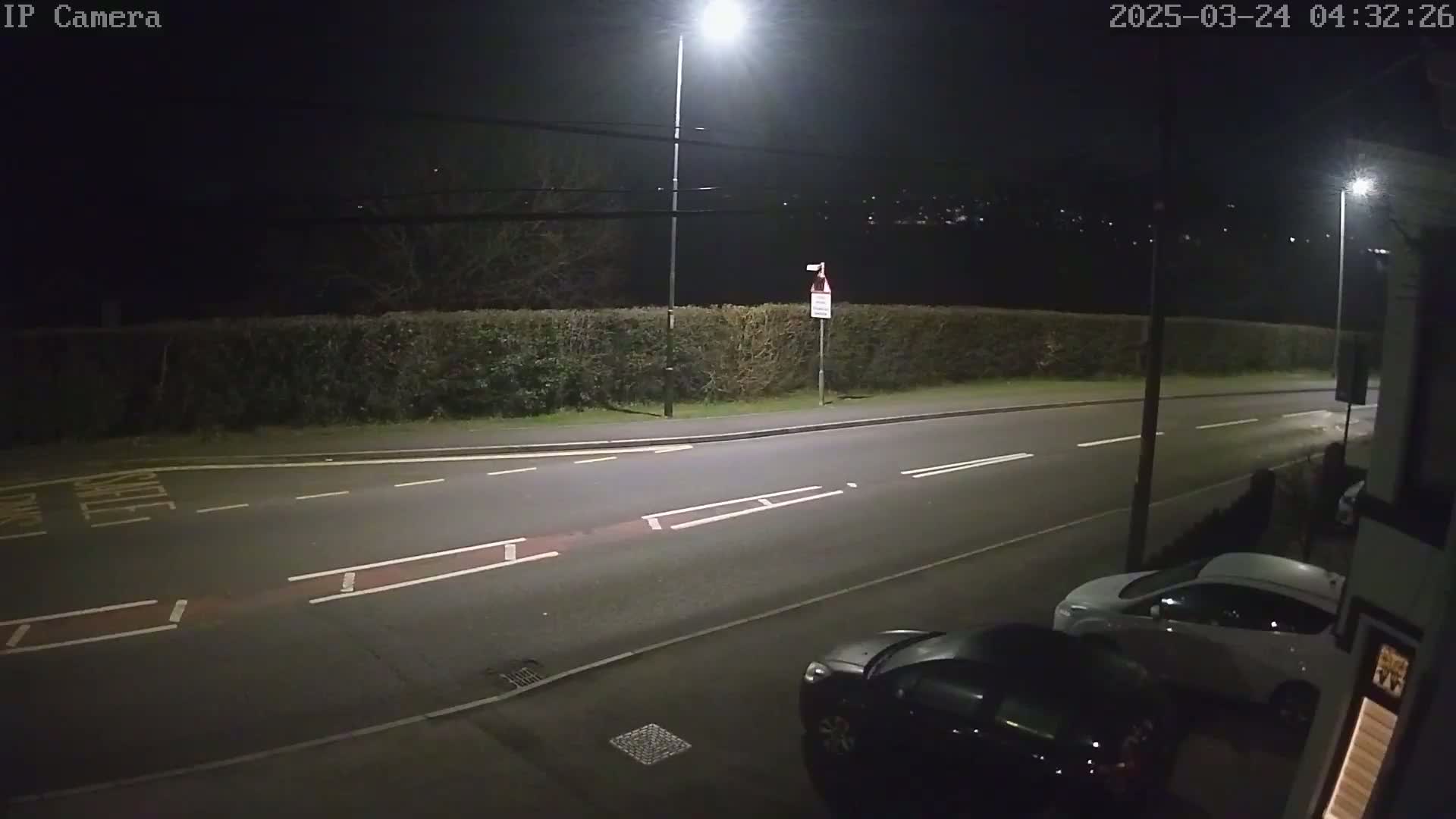 A nighttime street scene shows a roadway with two parked cars, illuminated by streetlights, bordered by a hedge and some trees under a dark sky.