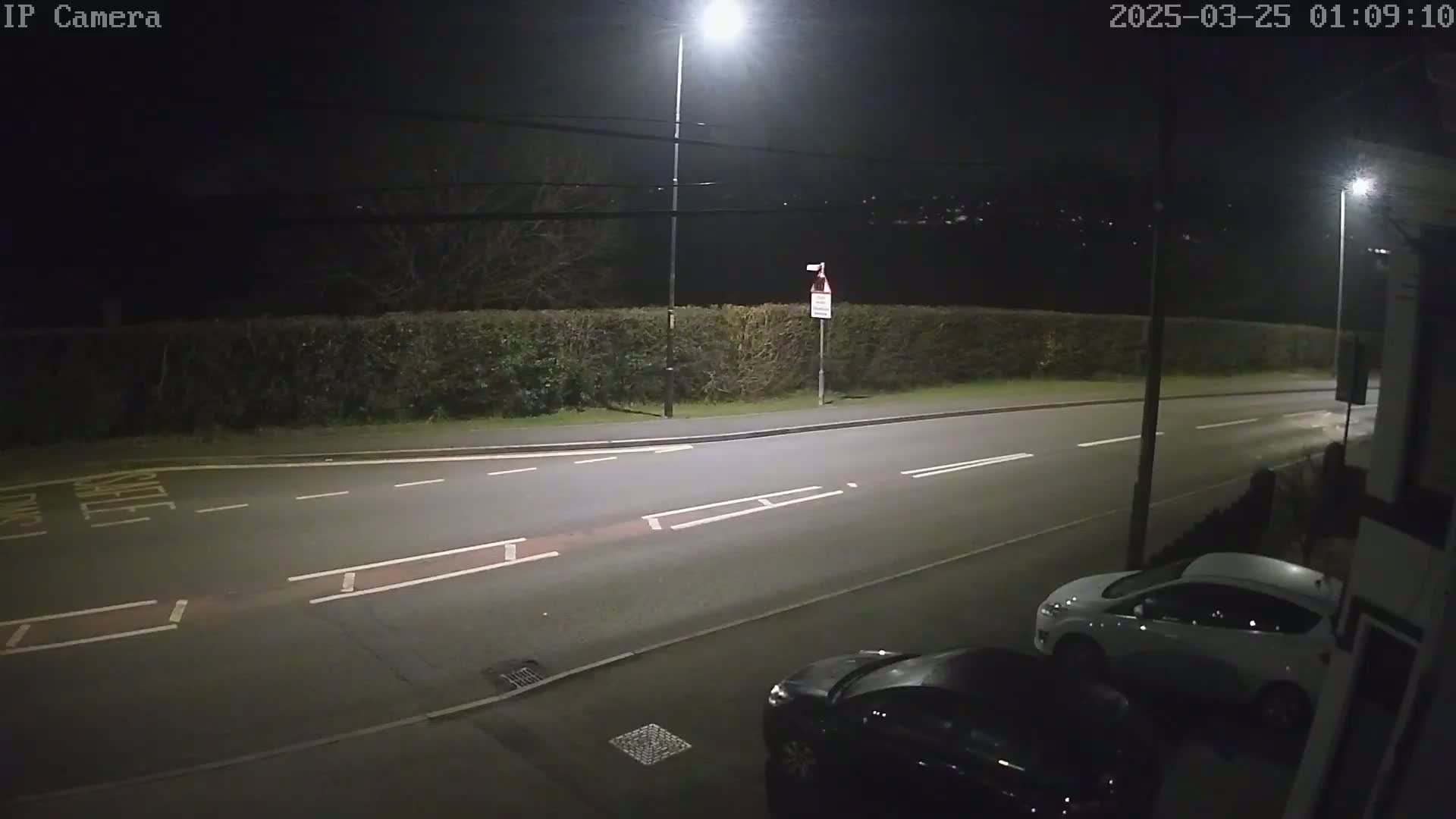 A nighttime street scene shows a two-lane road with parked cars, a hedge, and streetlights illuminating the area under a dark sky.