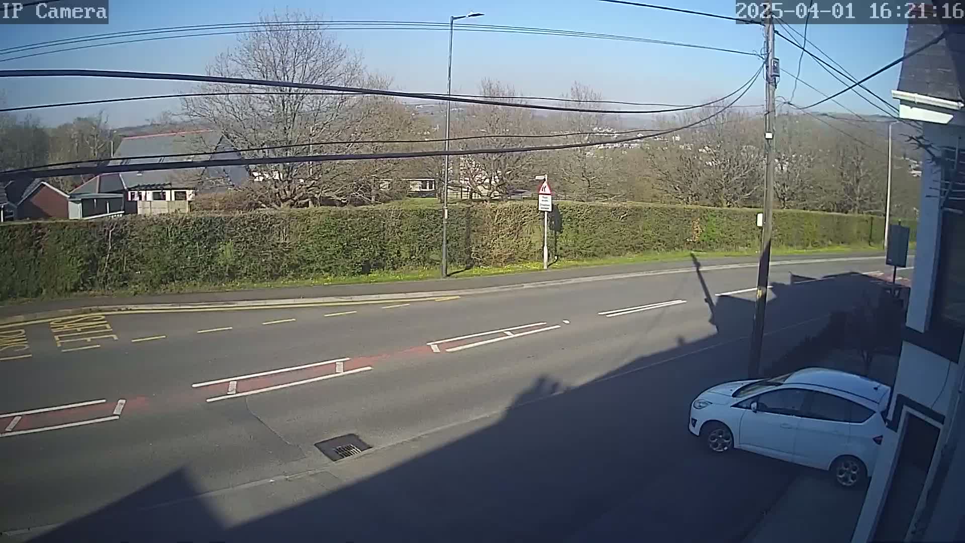 A sunny day shows a street scene with a white car parked at the roadside, across from a hedgerow, and a building in the distance.