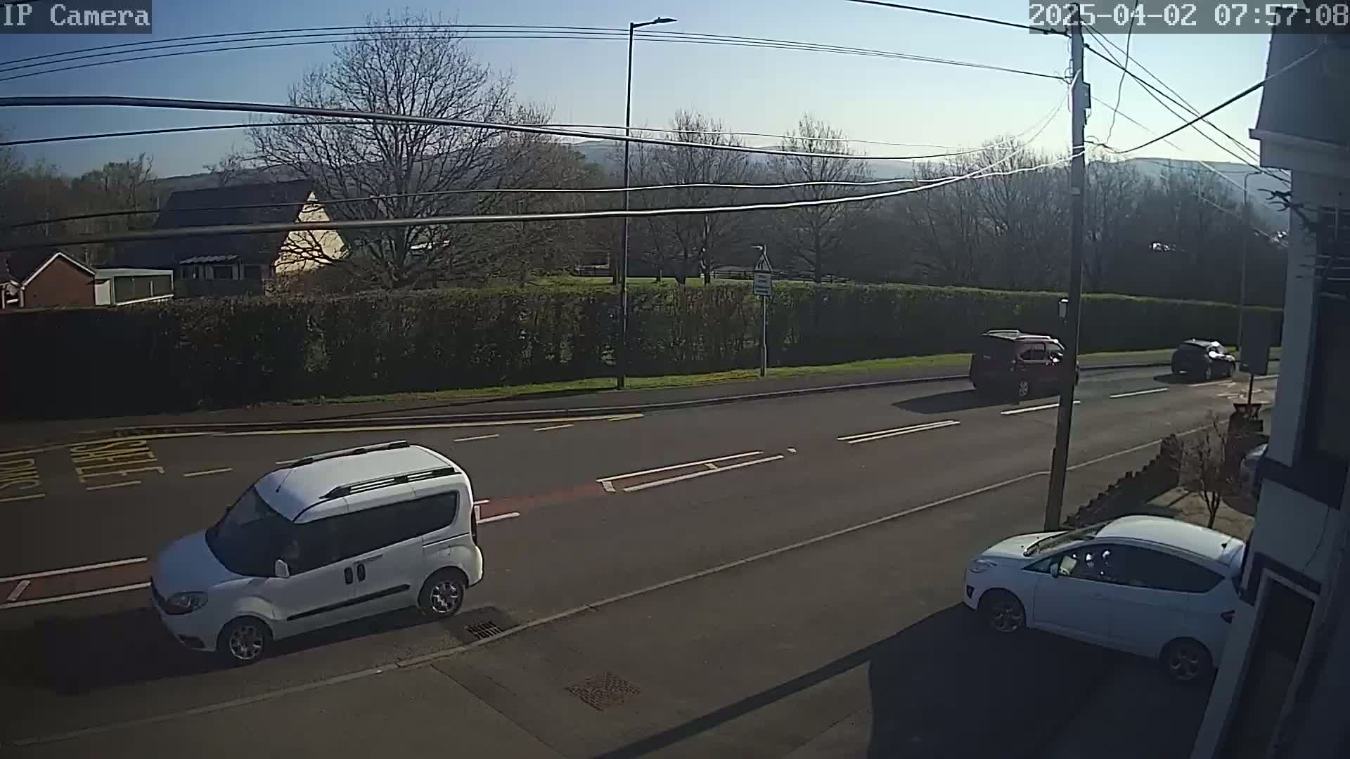 A sunny day shows a road with several cars, including a white van and a white hatchback parked, and a red SUV and dark-colored car driving.
