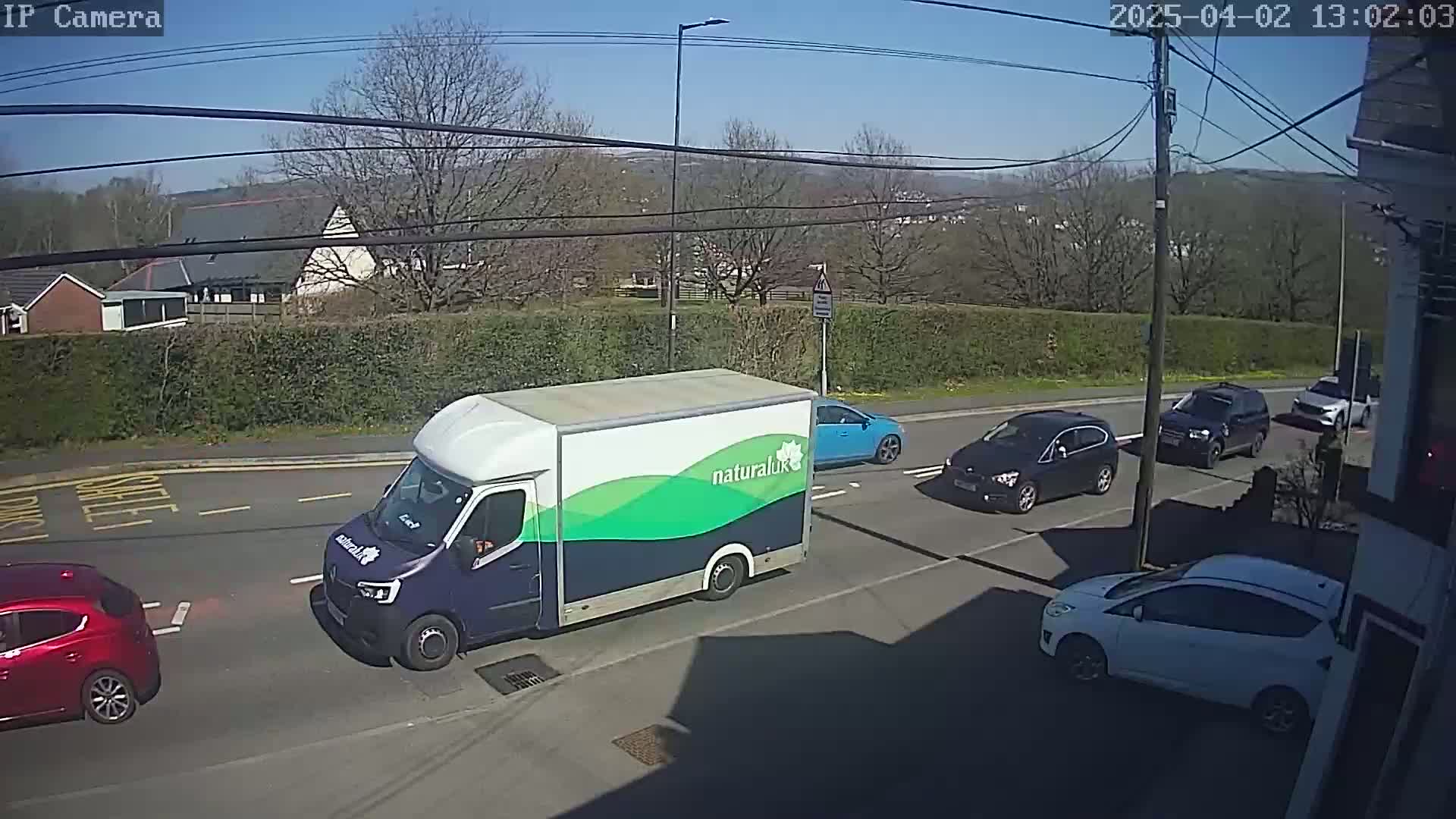 A blue and green delivery van is parked on a sunny street alongside several other cars.