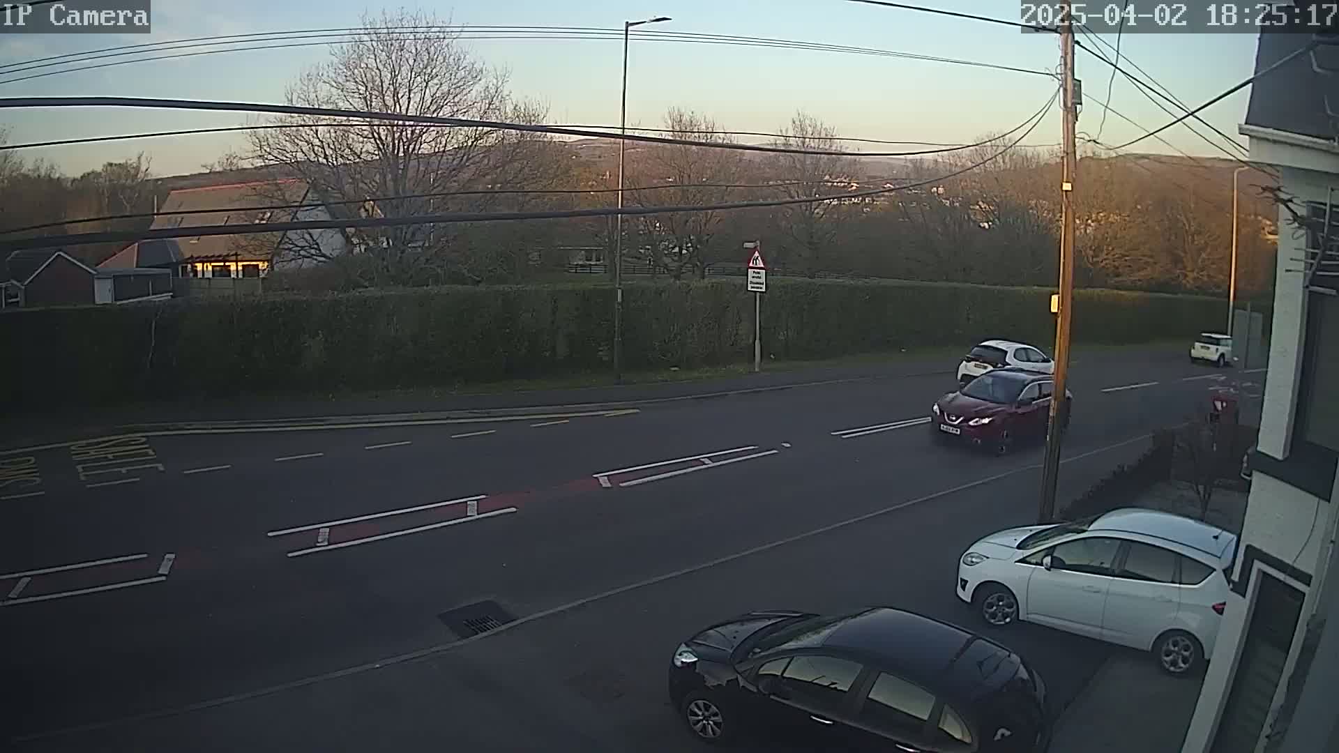 Several cars are parked and driving on a residential street at dusk, with a line of trees and houses in the background under a clear sky.