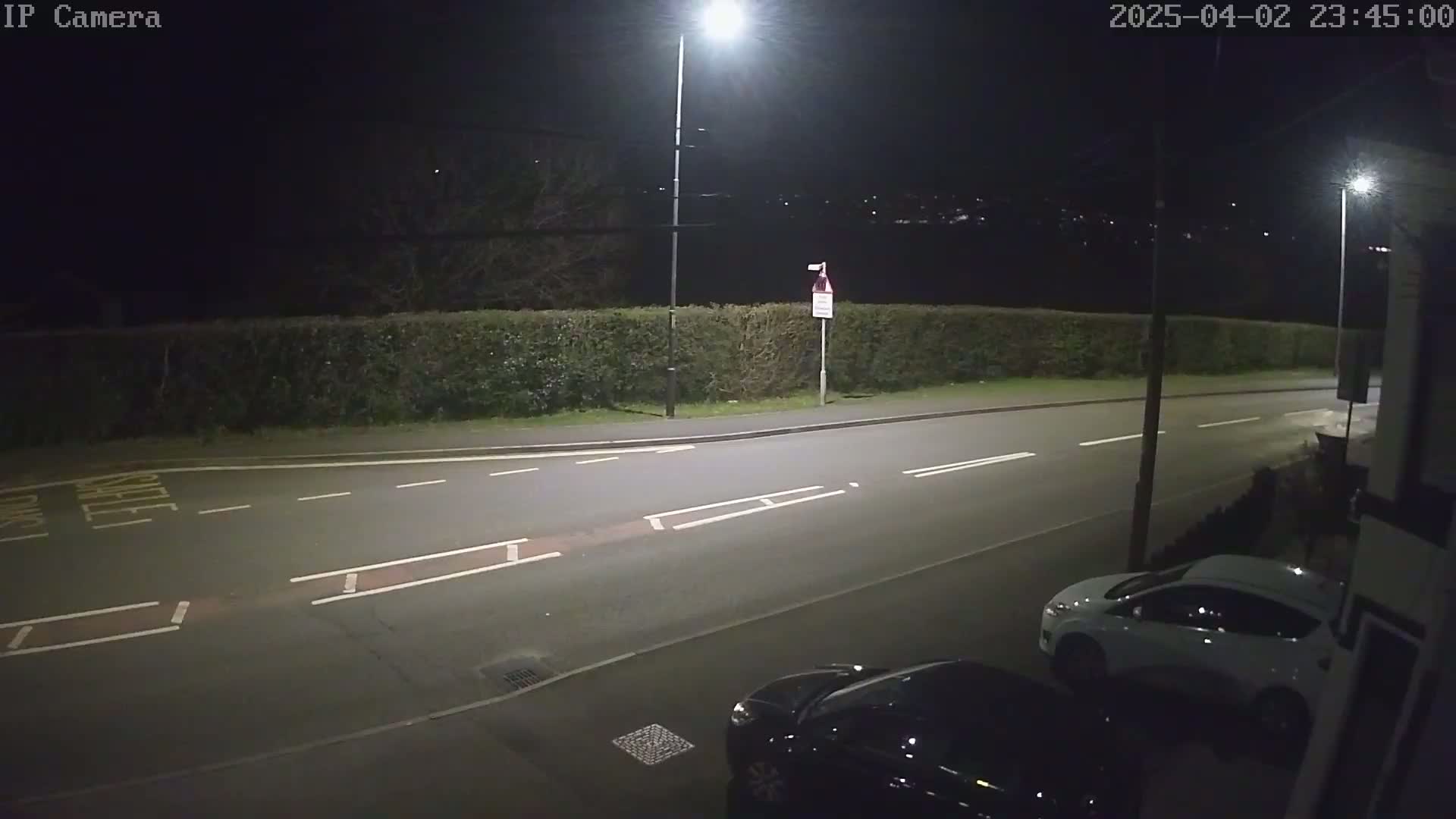 A nighttime street scene shows a road with parked cars, illuminated by streetlights, bordered by a hedge.
