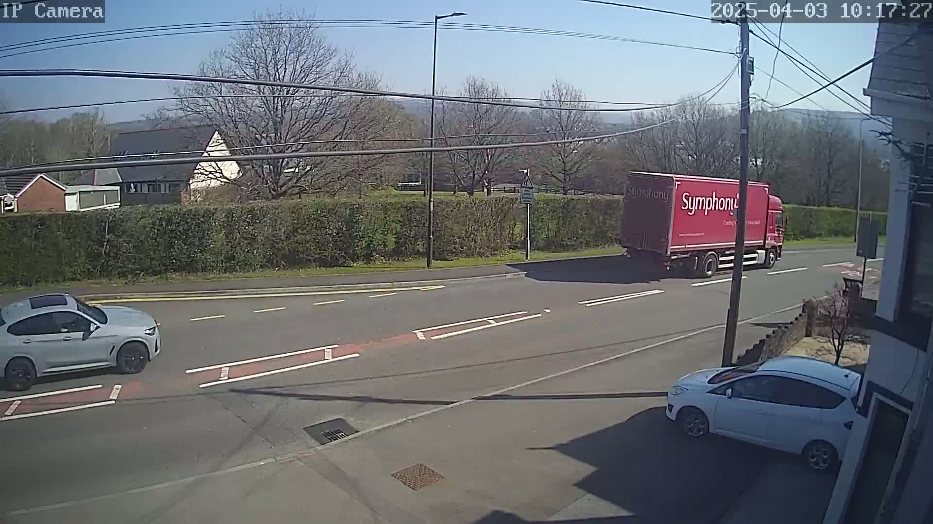 A sunny day shows a gray SUV and a white hatchback parked near a road where a red semi-truck is driving past.