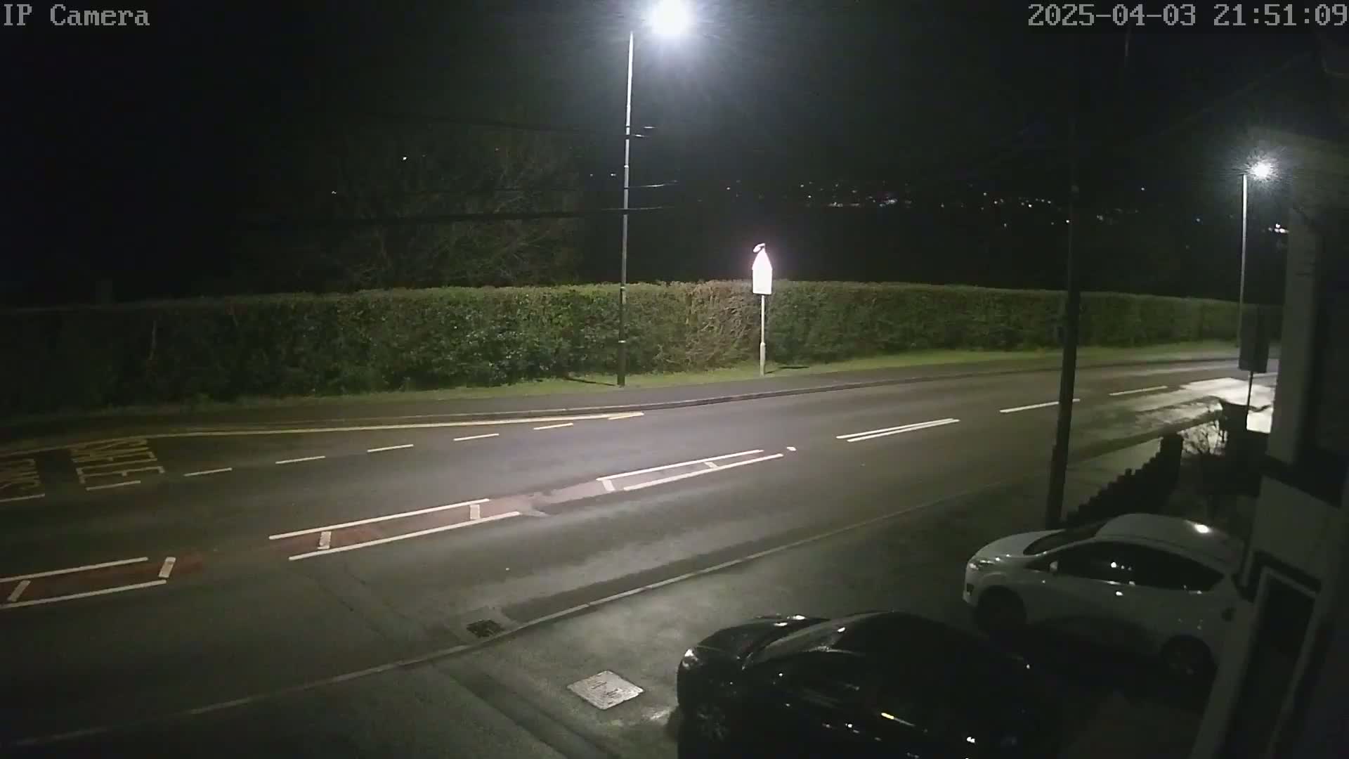 A nighttime street scene shows a road with parked cars, illuminated by streetlights, and bordered by a hedge.