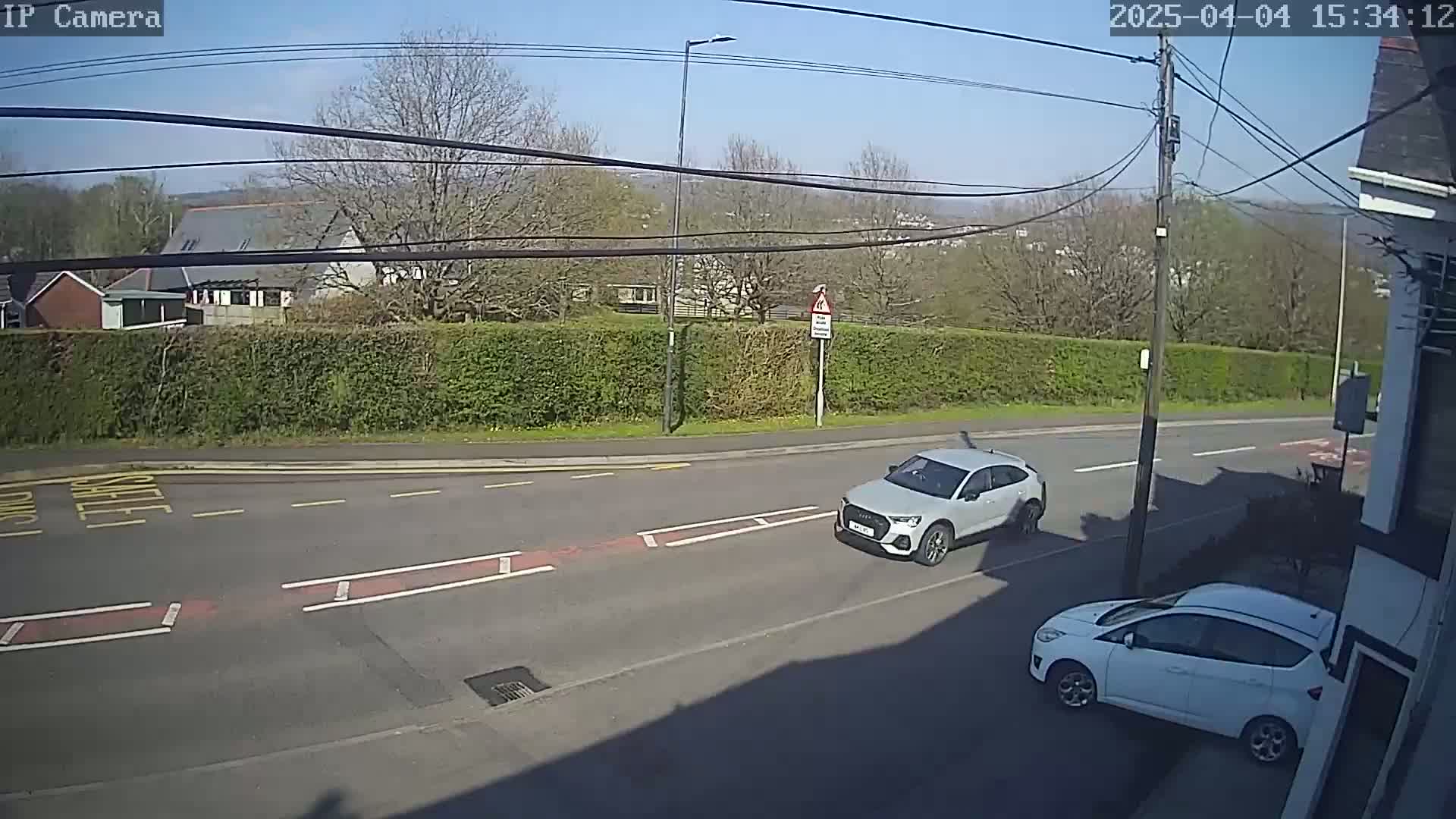 A sunny day shows two white cars on a street, one driving away and another parked near a building;  a hedge and some buildings are visible in the background.