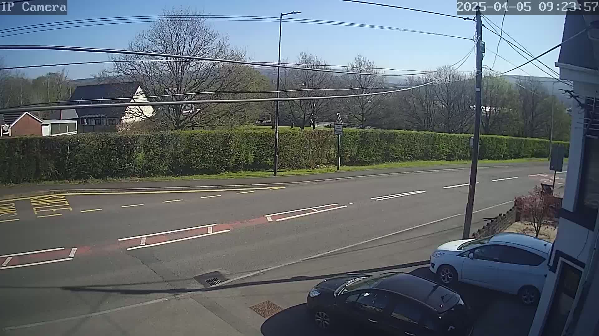 A sunny day shows a road with two parked cars, one white and one dark-colored, next to a hedgerow, with buildings and trees visible in the background.