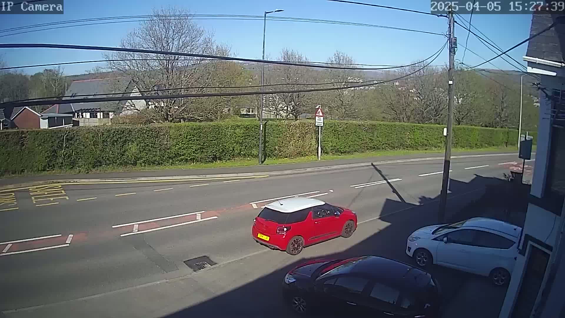 A sunny day reveals a road scene with a red car, a white car, and a black car partially visible, all parked near a hedge bordering a residential area.