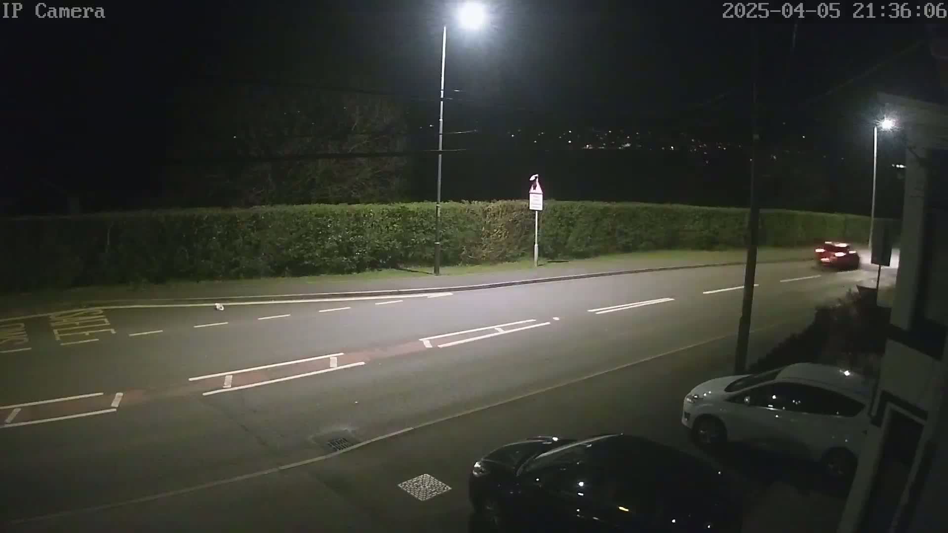 A nighttime street scene shows a dark-colored car parked near a white car, with a red car driving away in the distance, all under nighttime lighting conditions.