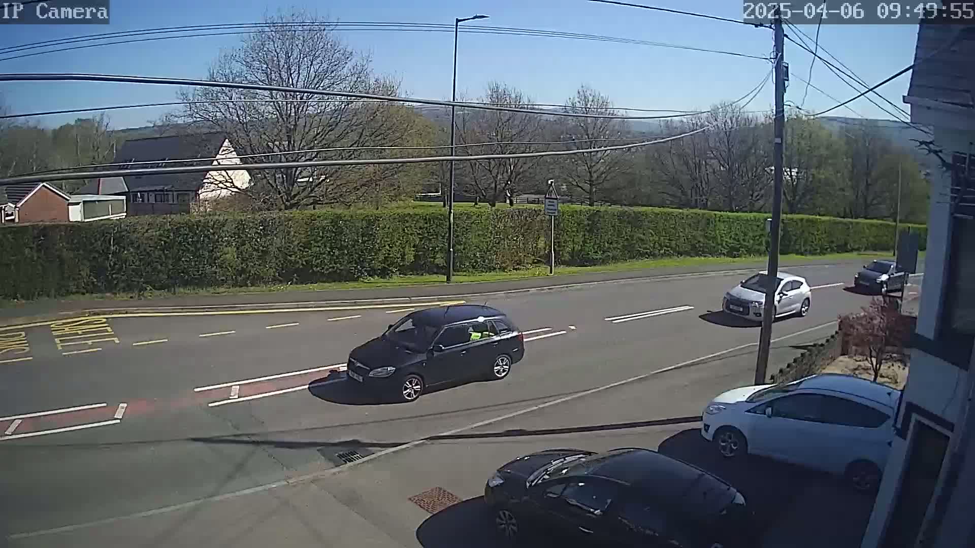 Several cars are driving on a road under a sunny sky, past a hedgerow and houses.