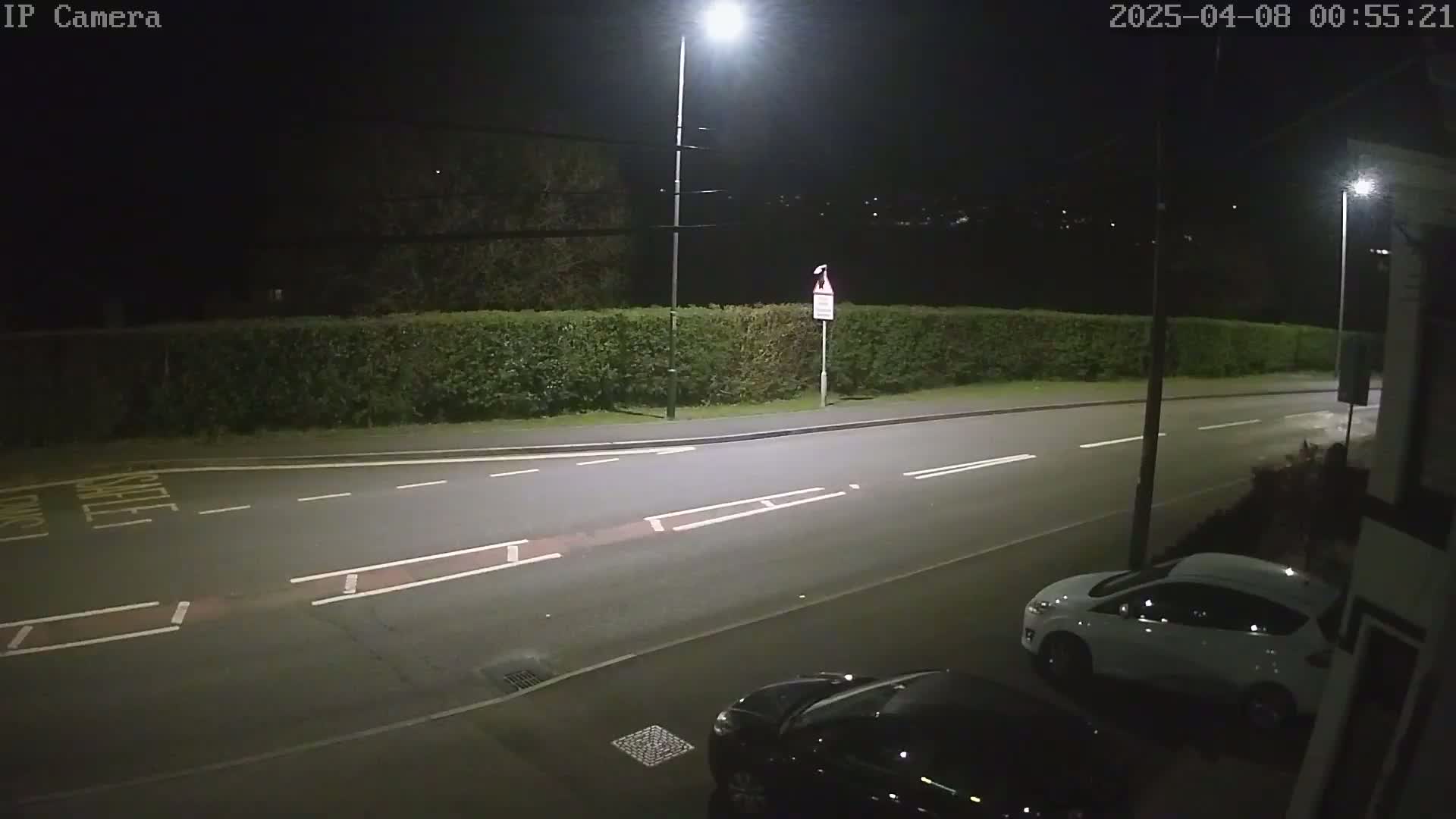 A nighttime street scene shows a road with two parked cars, a hedge, and streetlights illuminating the area.