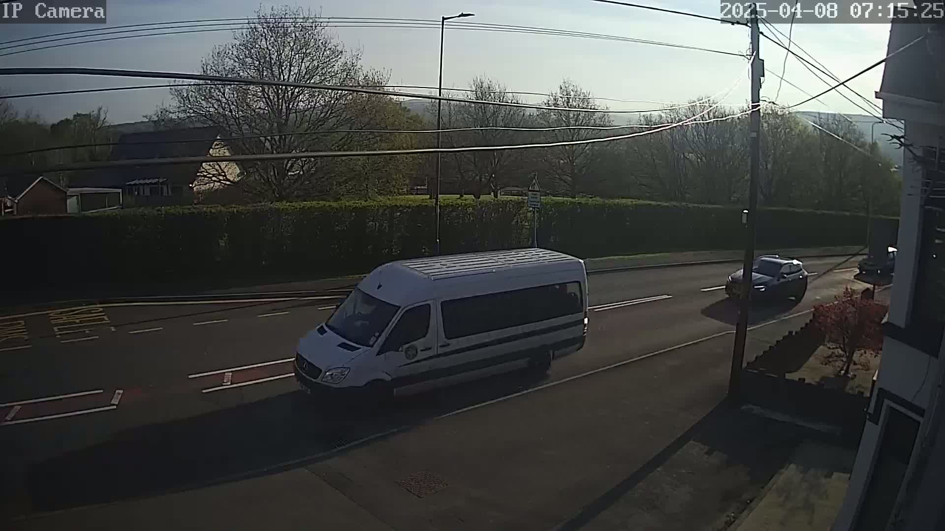 A white van and a dark-colored car are driving on a road bordered by a hedge and trees under a mostly sunny sky.