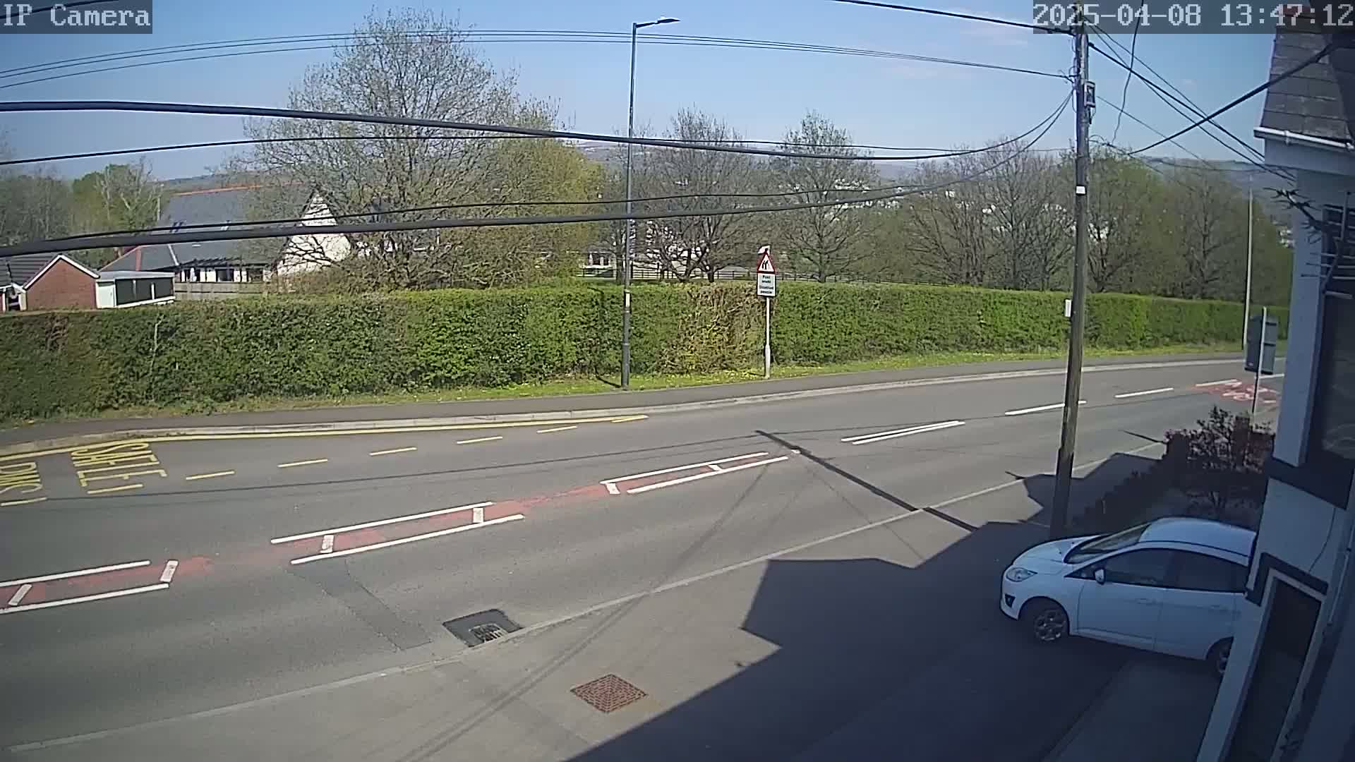 A sunny view of a residential street with a white car parked on the side, a tall hedge, and buildings in the background.