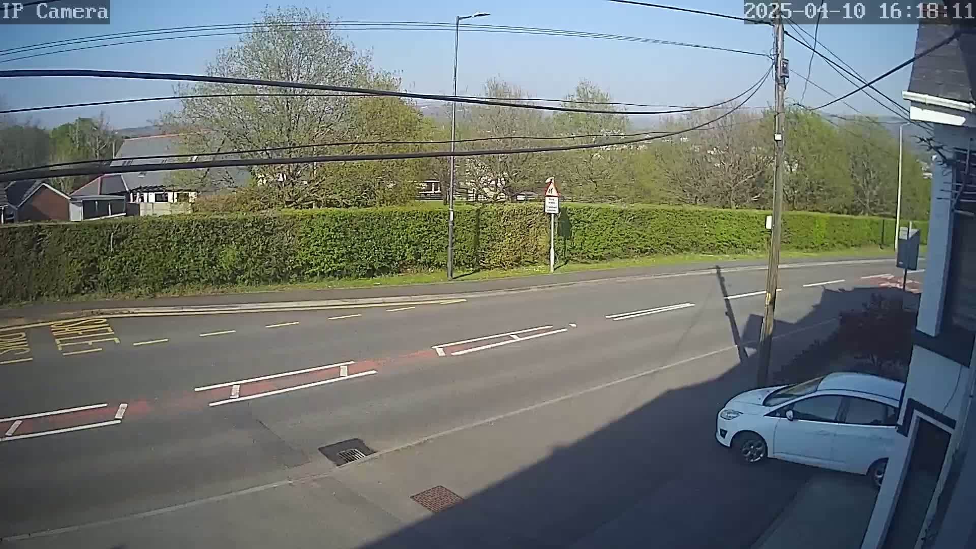 A sunny day reveals a white car parked on the side of a road, with a hedge and buildings in the background.