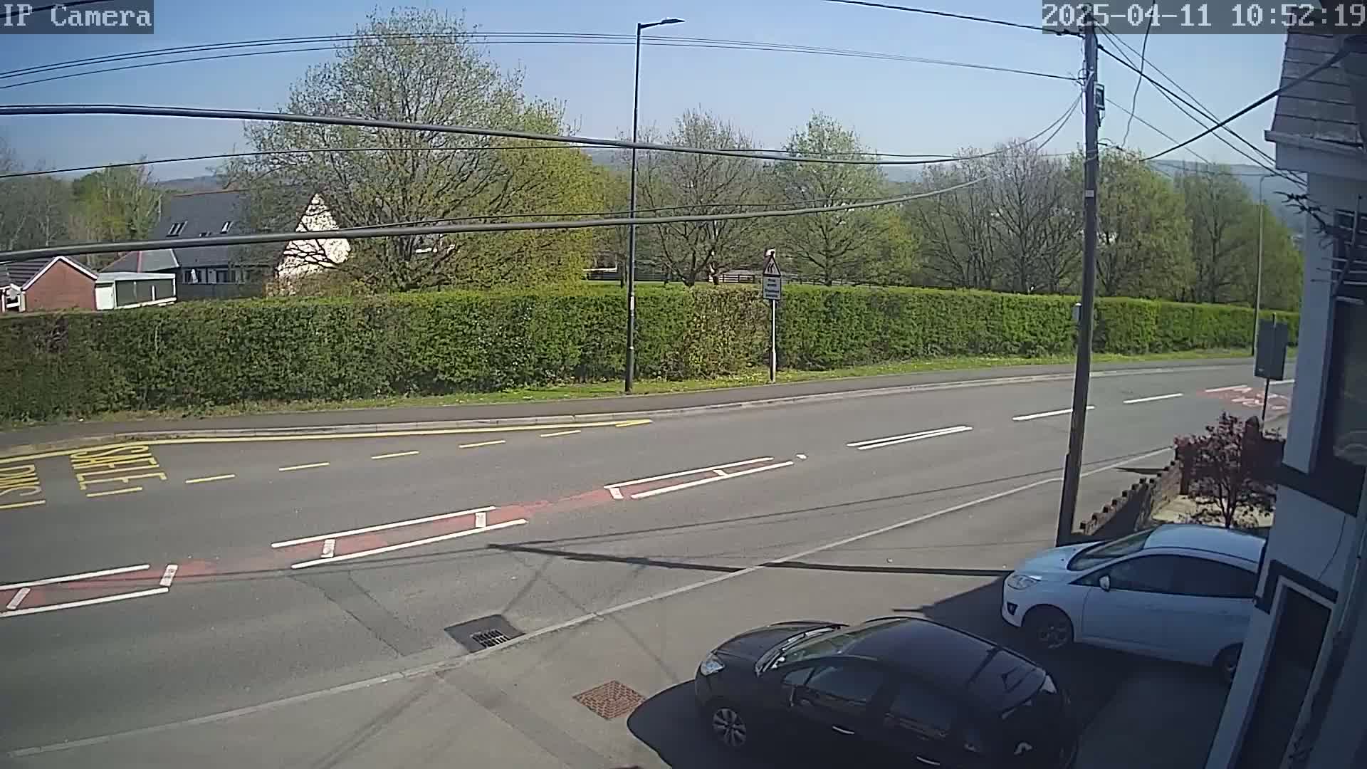 A sunny day reveals a residential street scene with two cars parked near a hedge, and a road with lane markings.