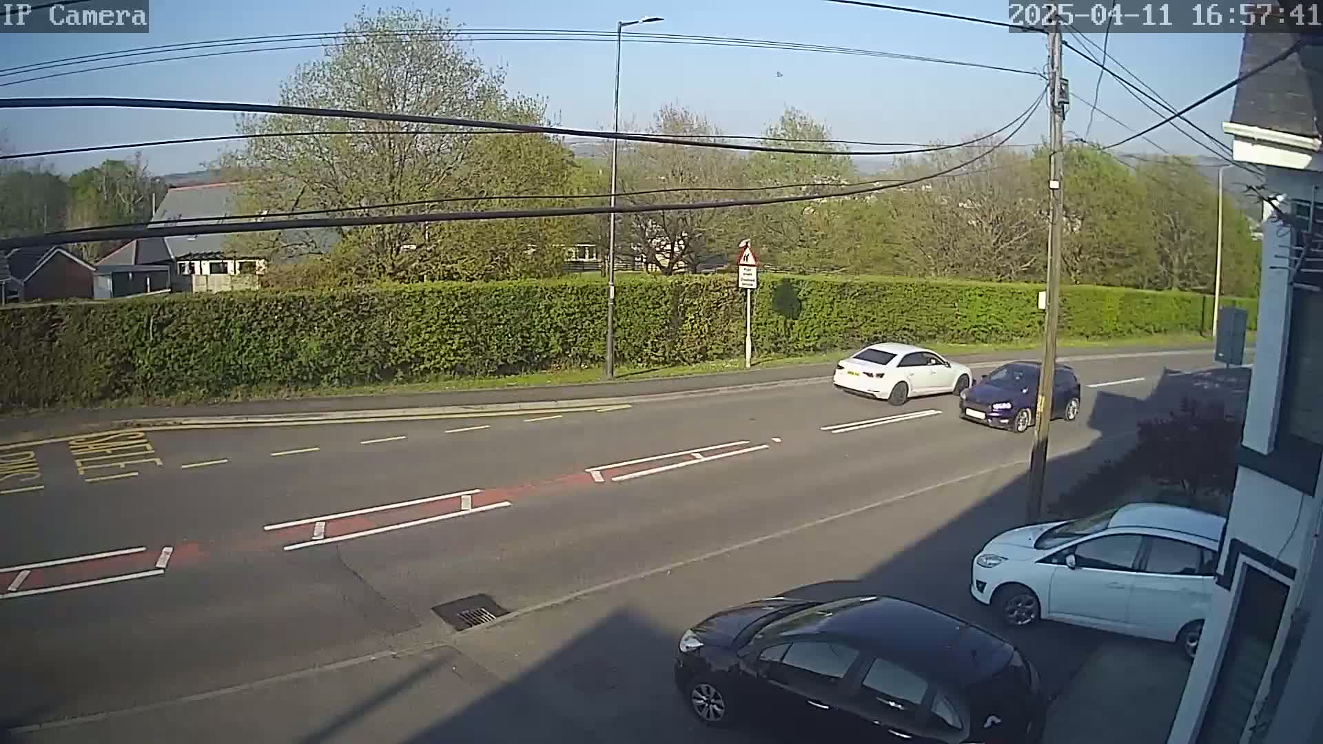 A sunny day shows three cars on a road bordered by a hedge, with additional cars parked nearby.