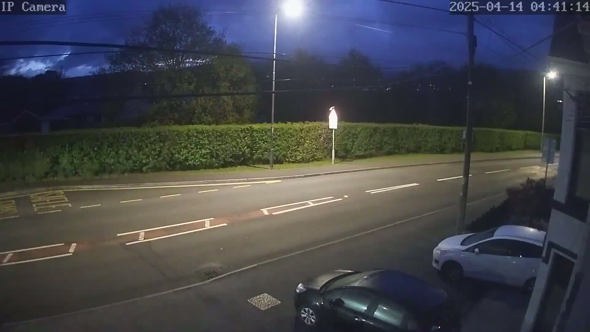 A dark, mostly clear night scene shows a road with two cars parked, a hedge, and a streetlight.
