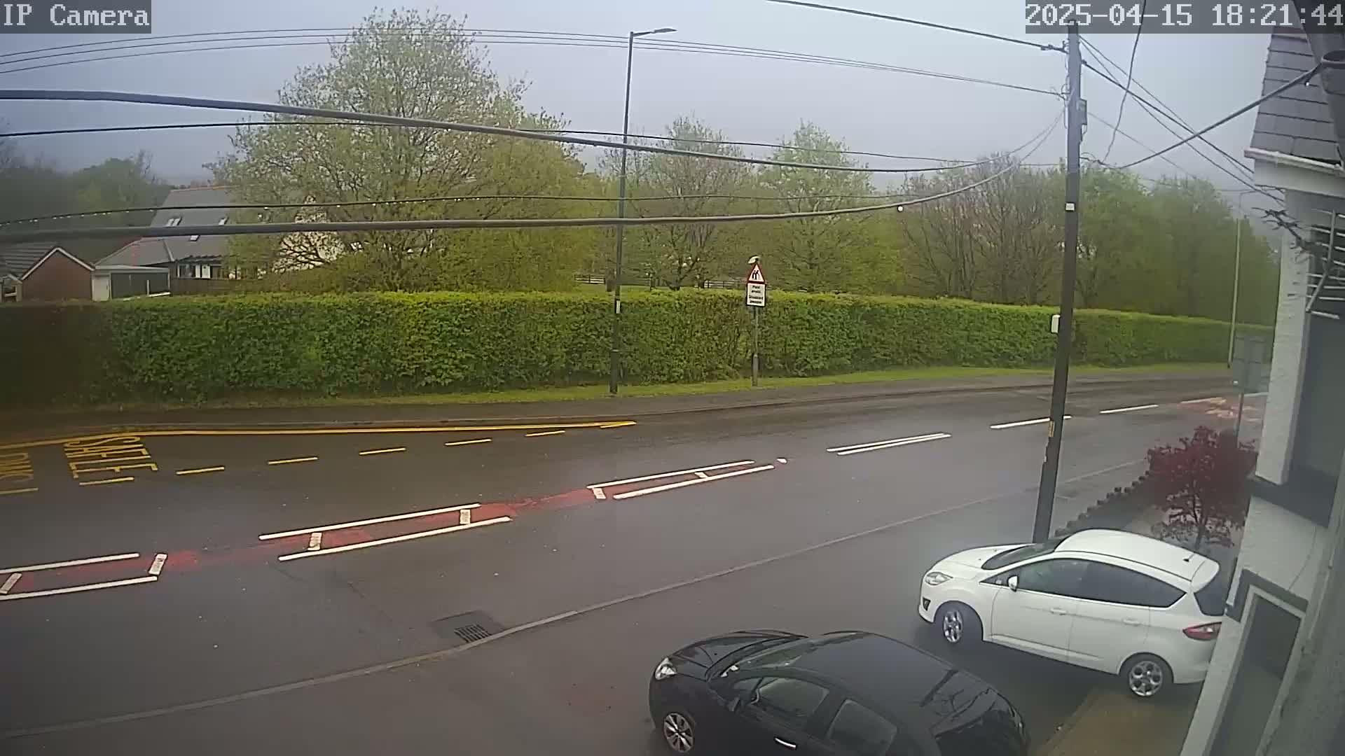 A wet road with two cars parked on the side, one white and one dark-colored, is viewed from a slightly elevated angle on an overcast day.