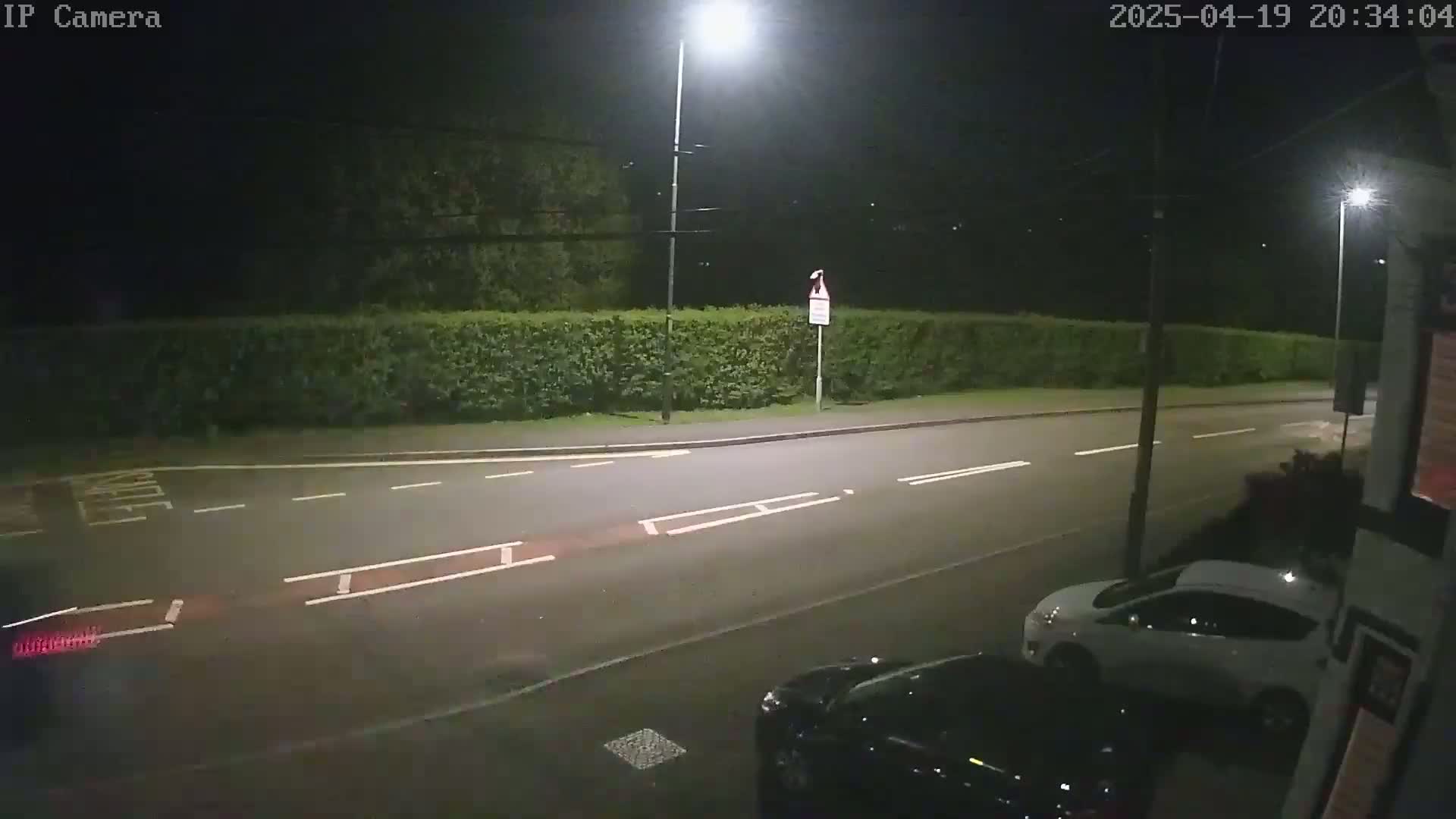 A nighttime view of a mostly empty road with a hedge and parked cars, lit by streetlights.