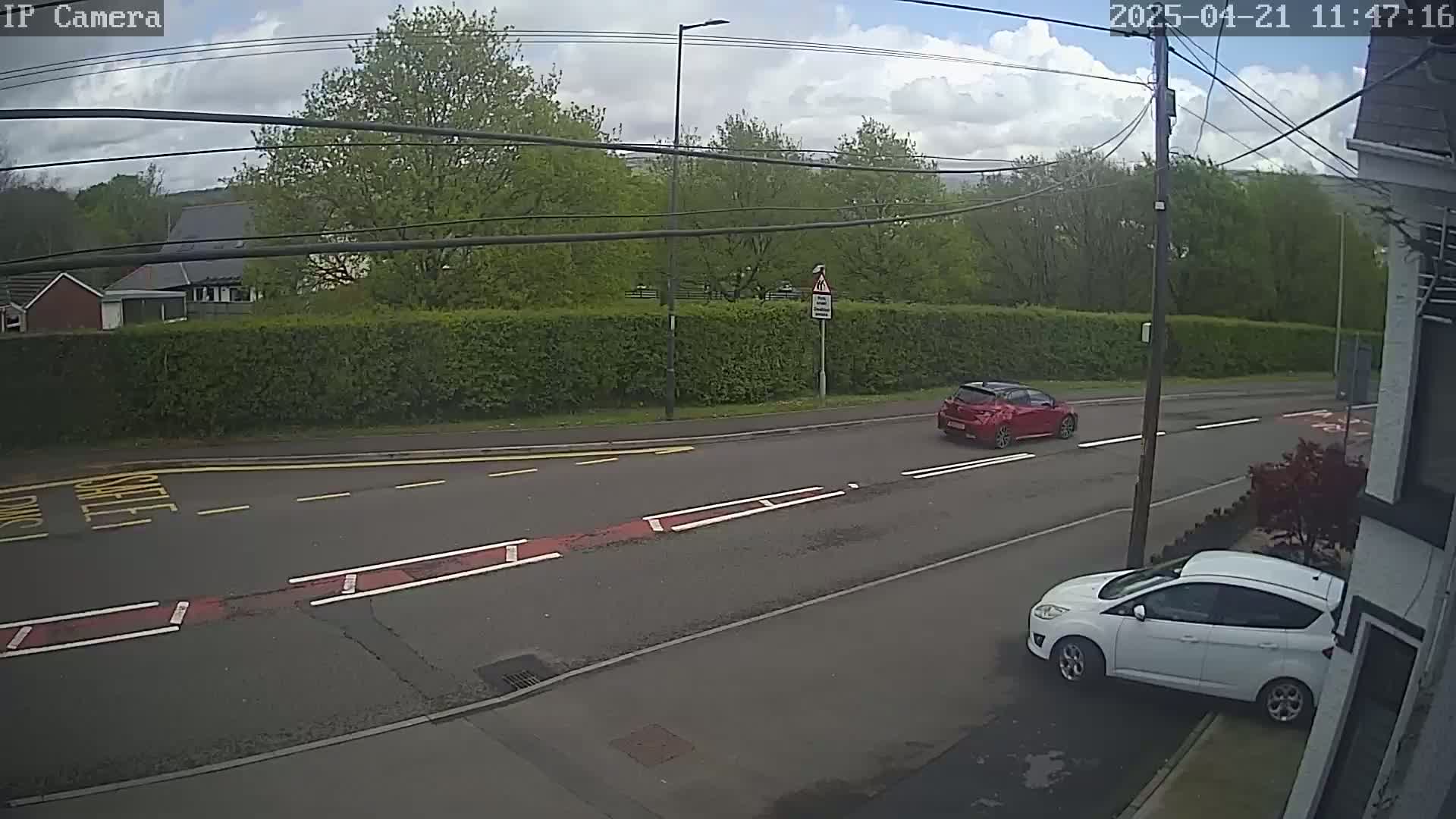 A partly cloudy day shows a red car driving down a residential street past a white car parked on the side, with a tall hedge and trees in the background.