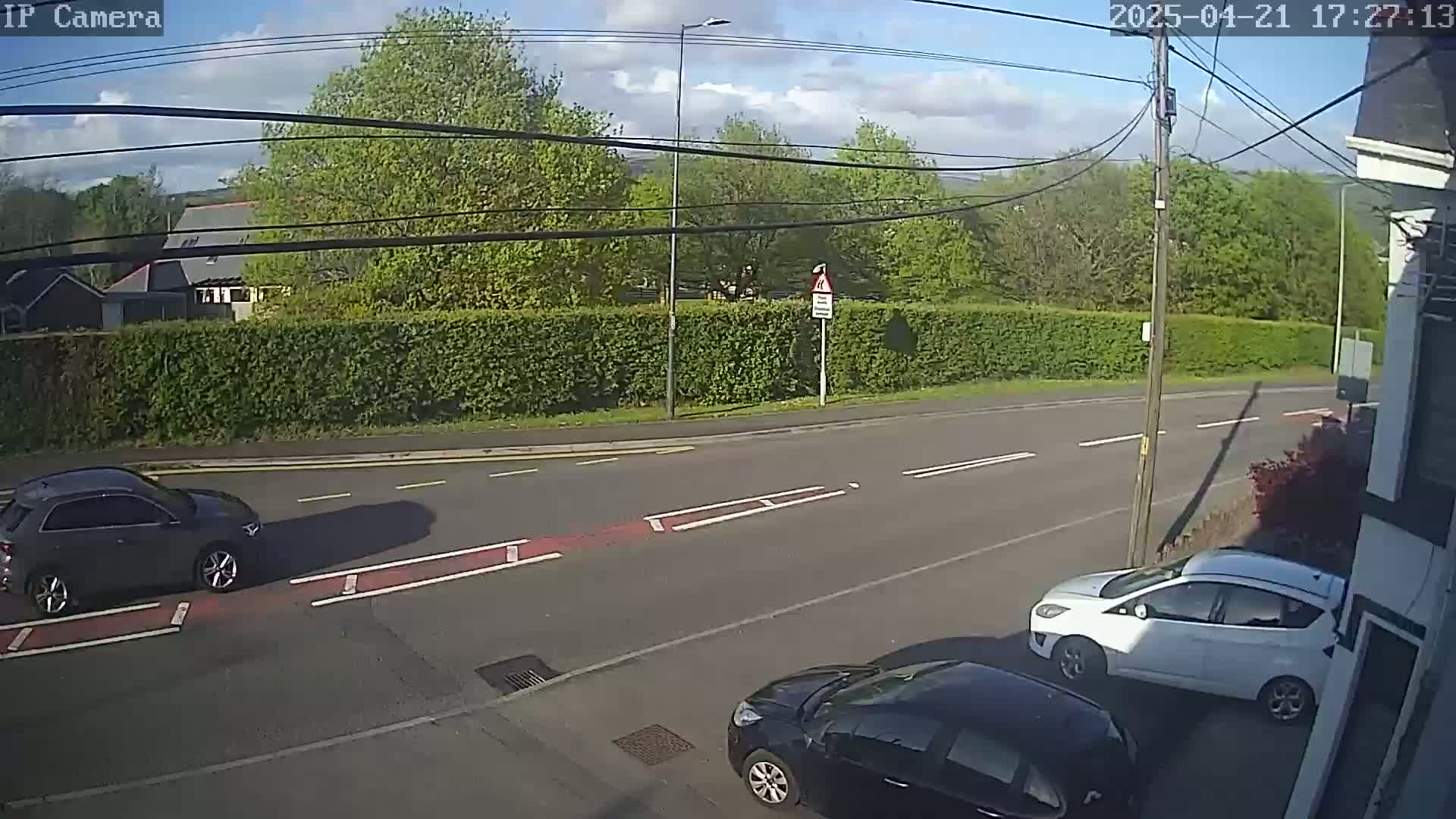 A partly sunny day shows three cars parked on a road beside a tall hedge, with more trees and buildings visible in the background.