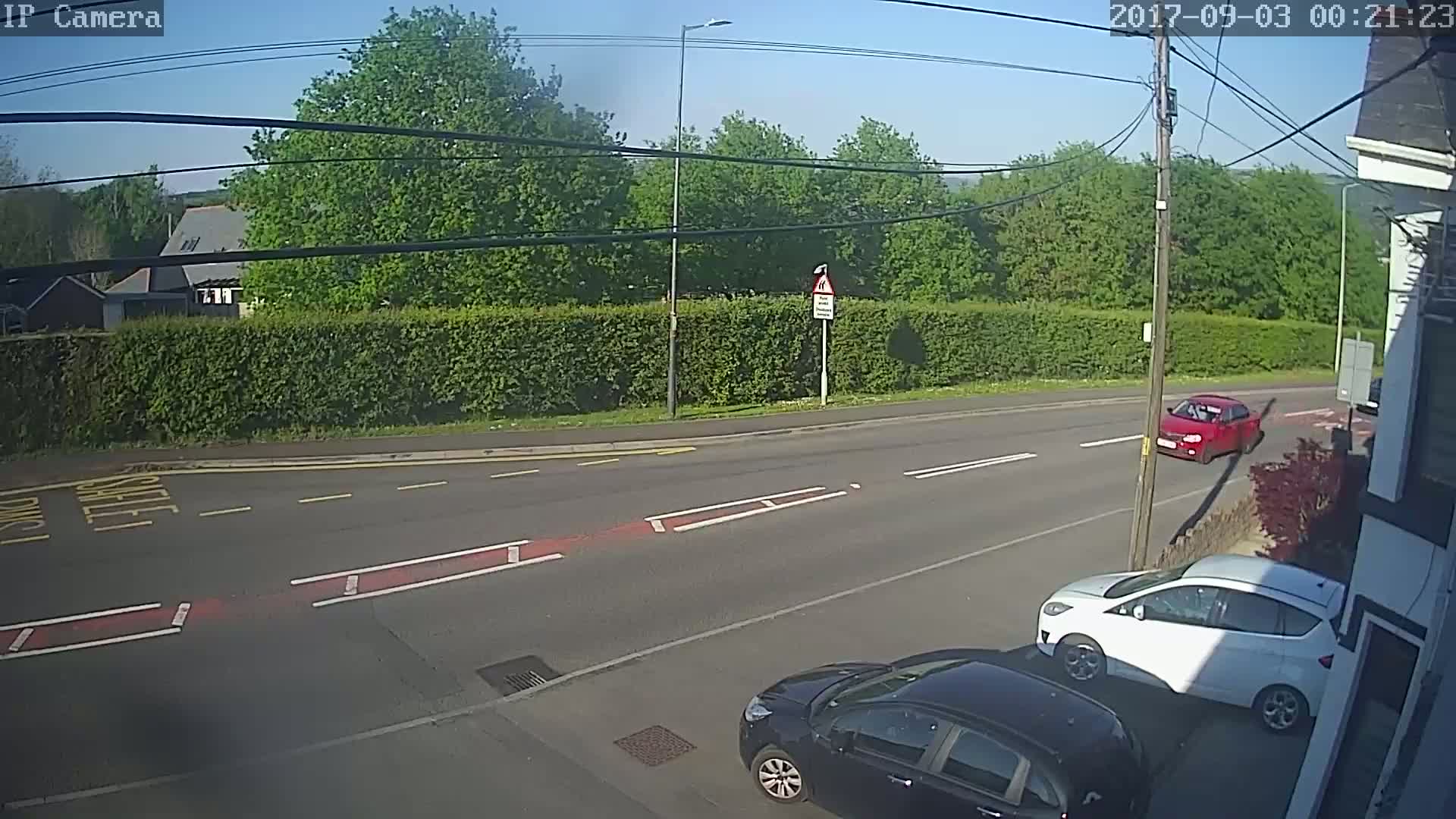 A sunny day shows a road intersection with three cars, two parked and one driving, and a hedgerow separating the road from a line of trees and buildings.