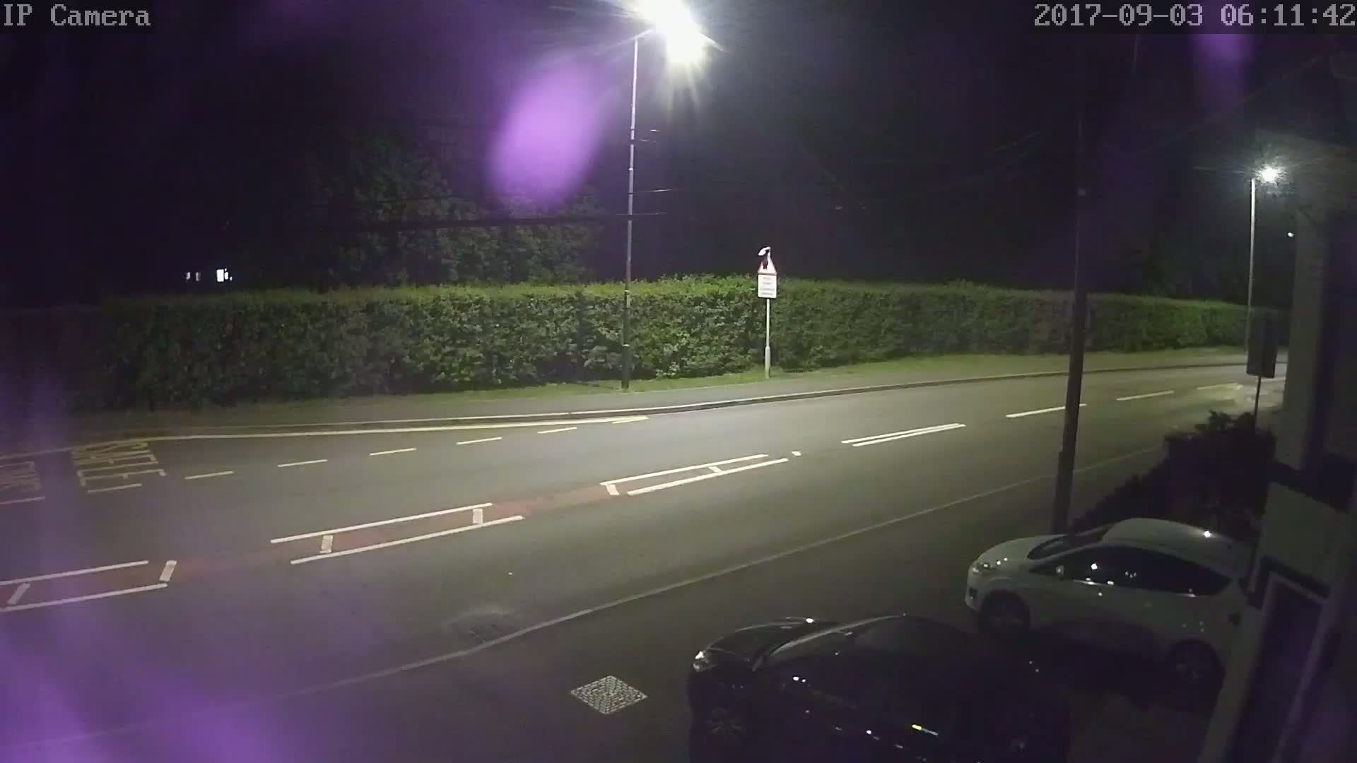 A nighttime street scene shows a road with white lane markings, a hedge, and two parked cars illuminated by streetlights.
