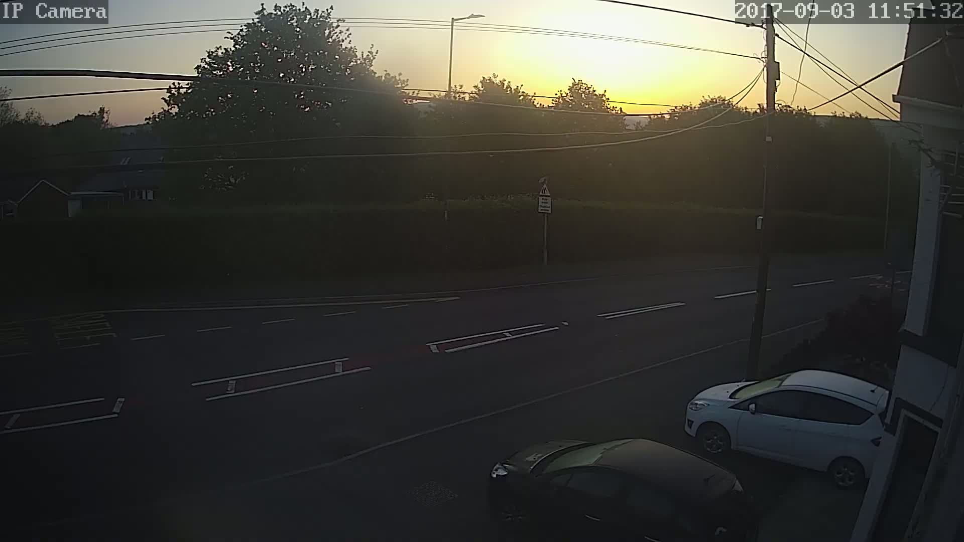 A sunrise illuminates a road with two cars parked near a building,  trees and shrubs line the road in the background.