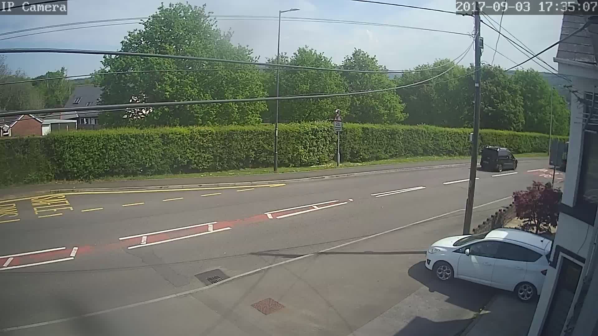 A sunny day shows a residential street with a white car parked near a building and a dark-colored van driving away on a road lined with a tall hedge.