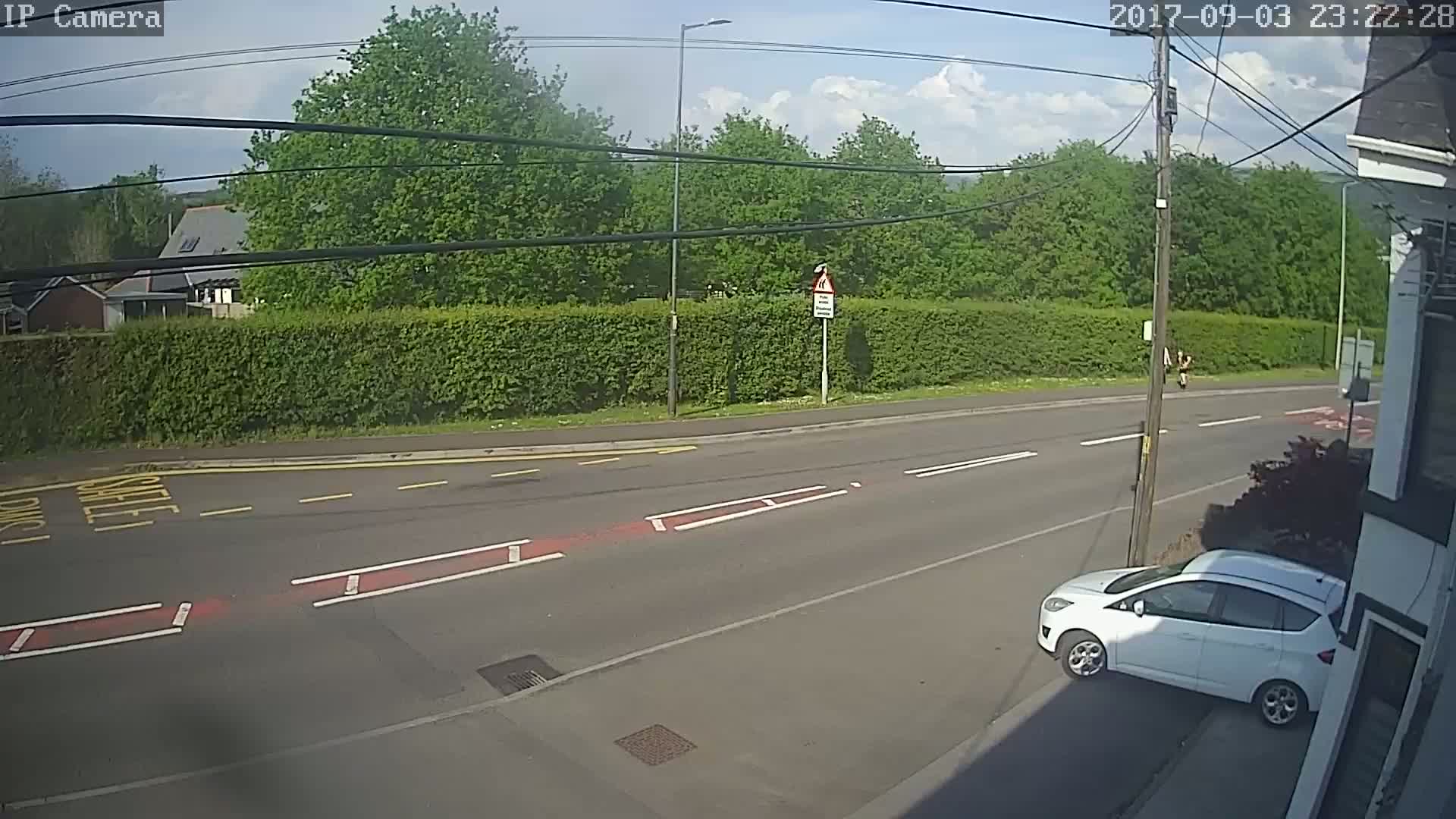 A mostly sunny day shows a white car parked on the side of a road that intersects with another road, bordered by a hedge and trees.