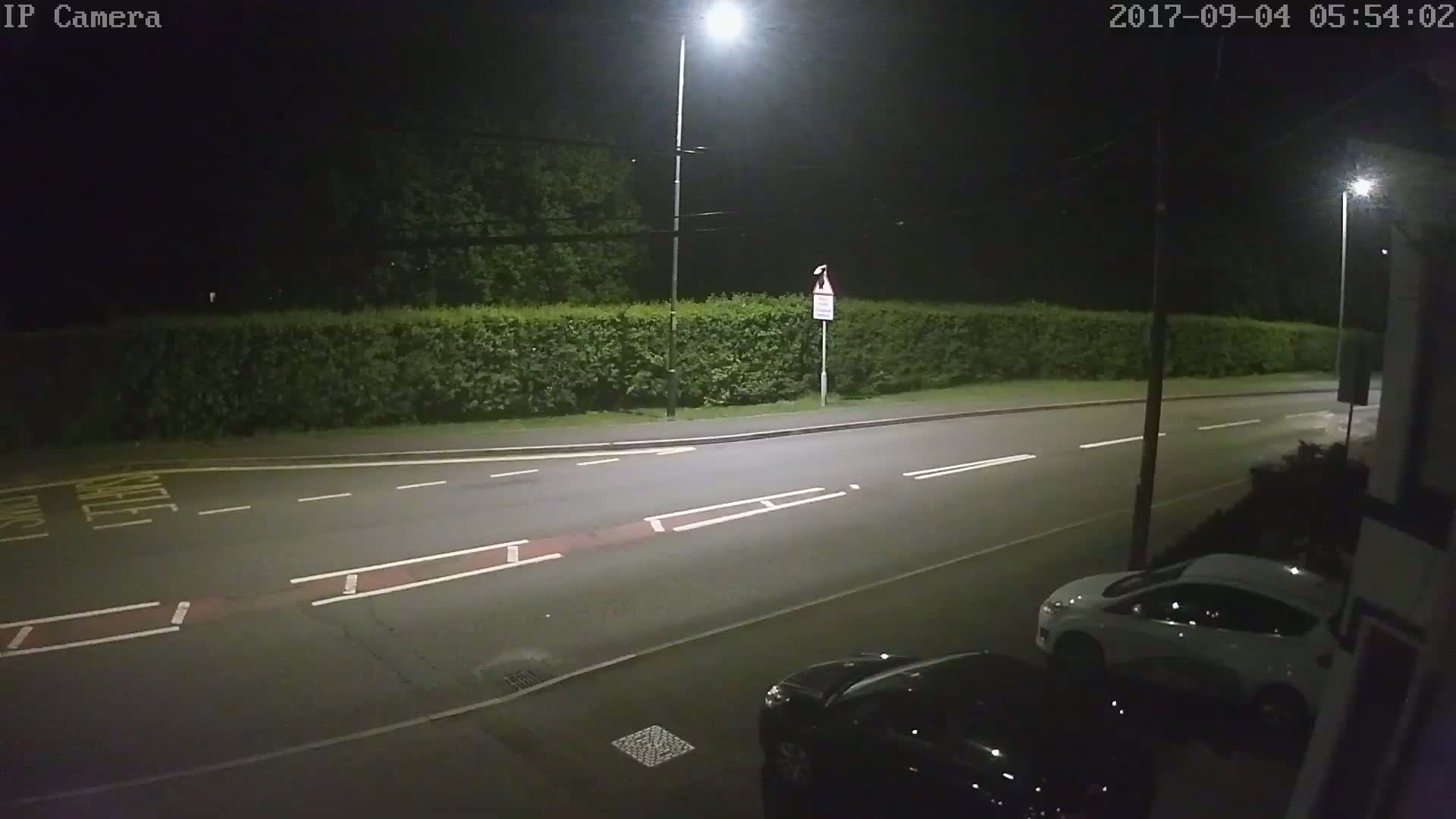 A nighttime street scene shows a road with white lane markings, a tall hedge, streetlights, and two parked cars under dark, clear skies.