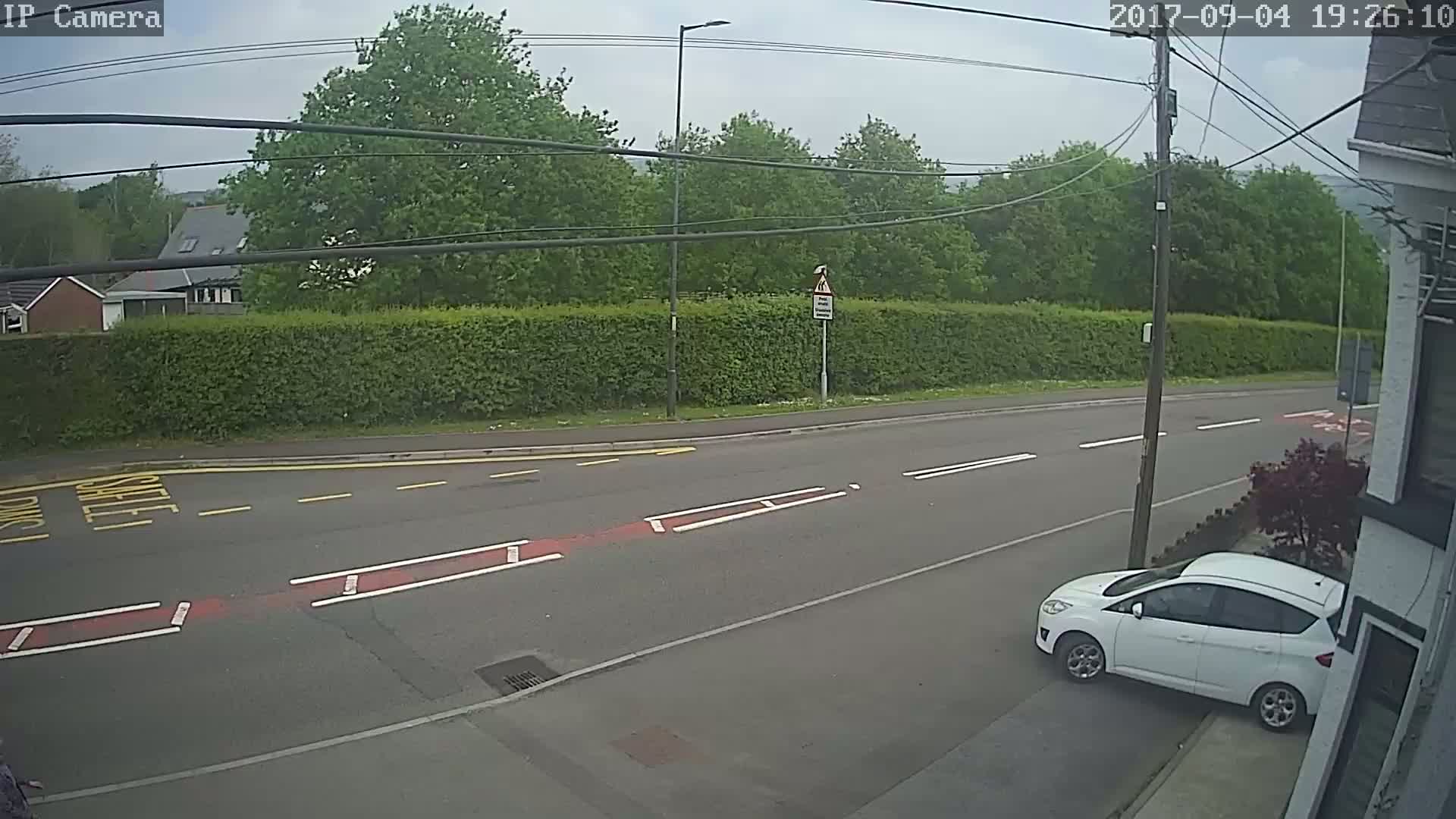 A white car is parked on the side of a road bordered by a tall hedge and trees under an overcast sky.