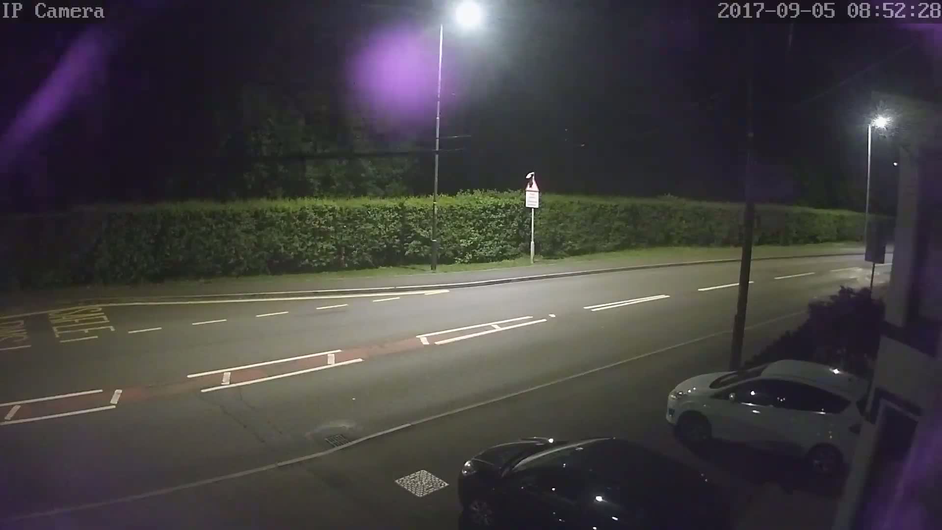 A nighttime street scene shows a road with parked cars under artificial light, next to a hedge.