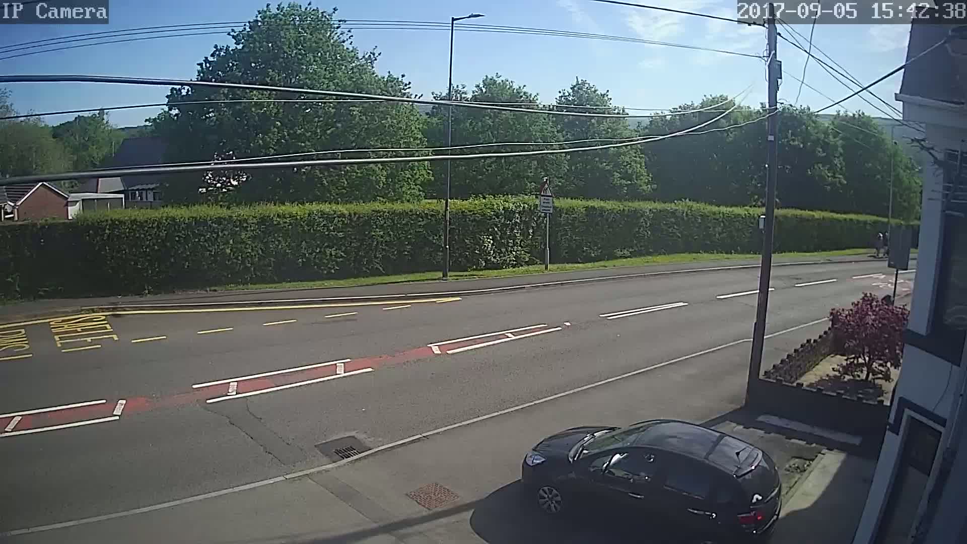 A sunny day shows a black car parked near a building beside a road with a hedge and trees in the background.