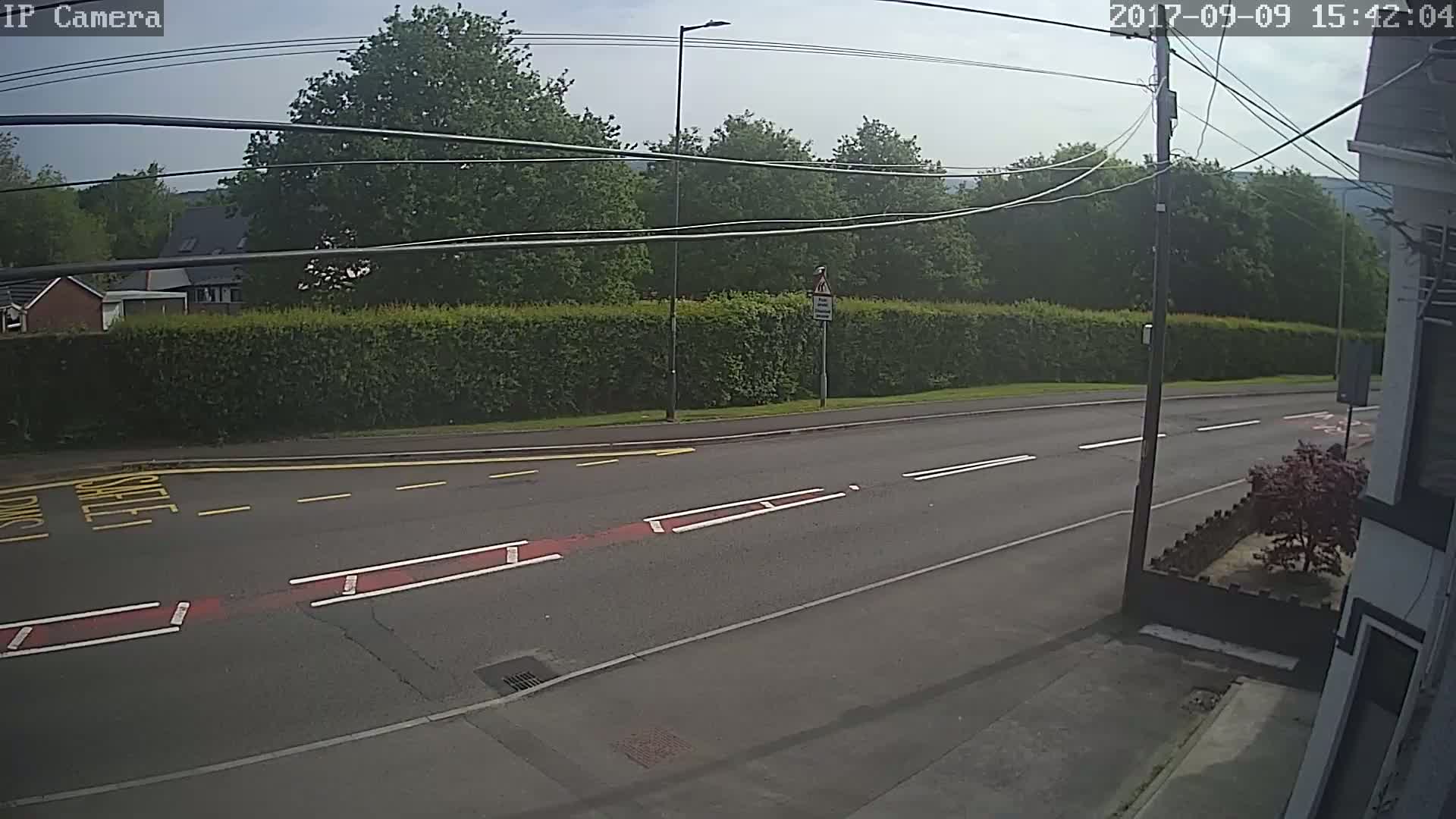 A mostly sunny day shows a roadway with yellow and red markings, a hedge, and trees beyond it.