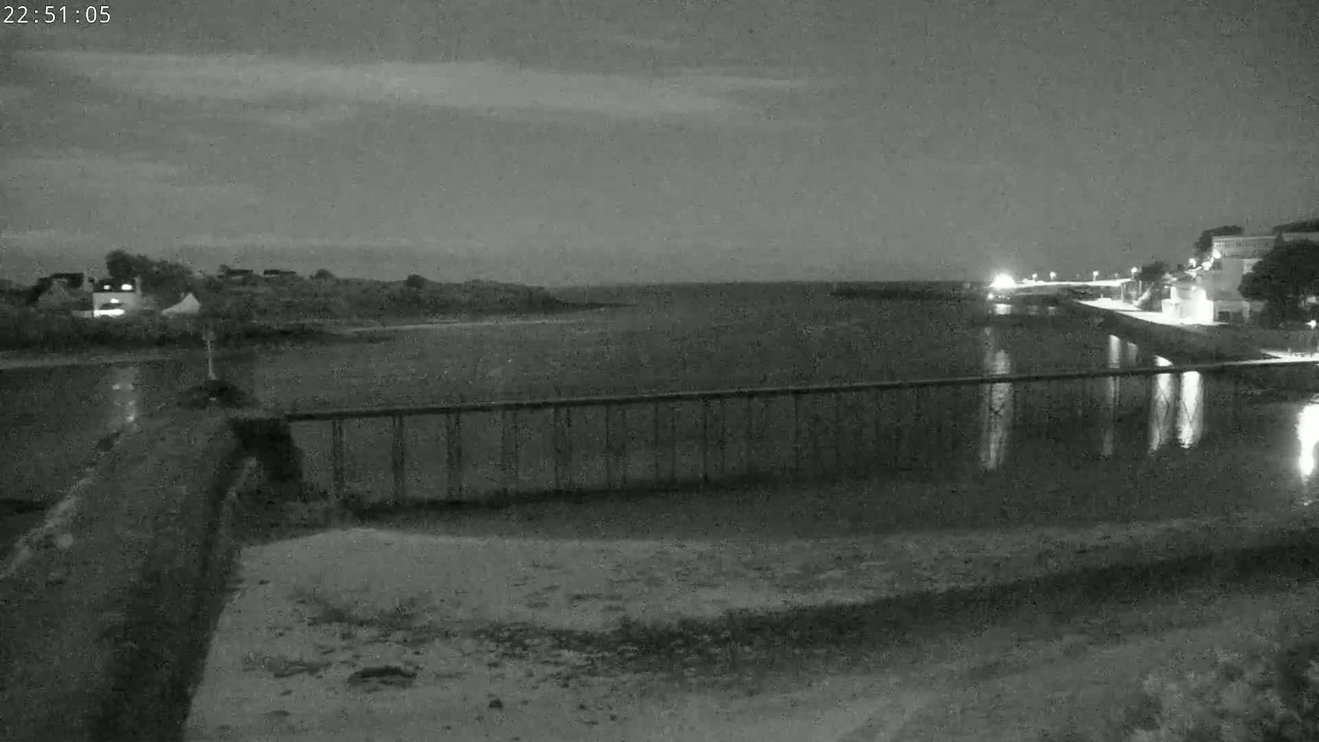A nighttime view shows a calm body of water under a partly cloudy sky, seen from behind a low metal railing.