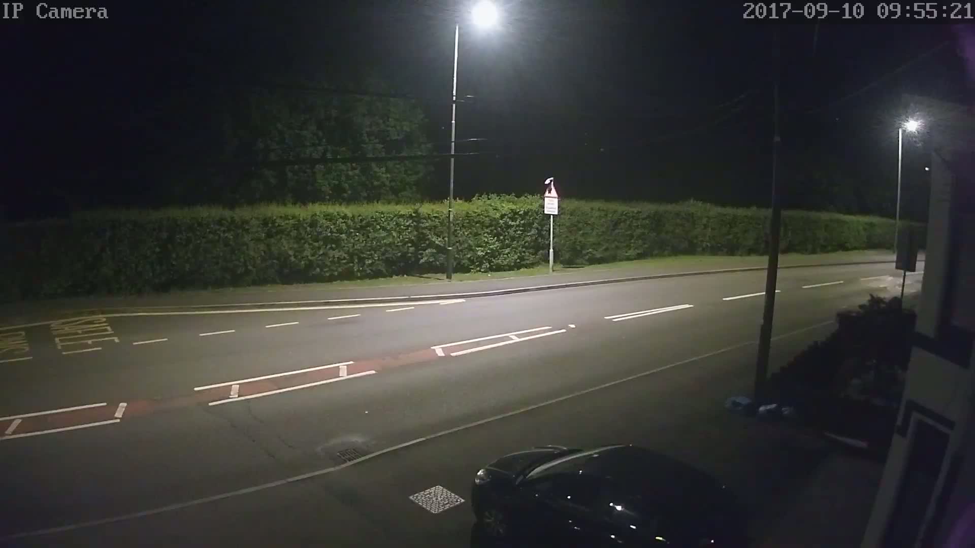 A nighttime view of a dark street illuminated by streetlights, showing a vehicle parked near a hedge.