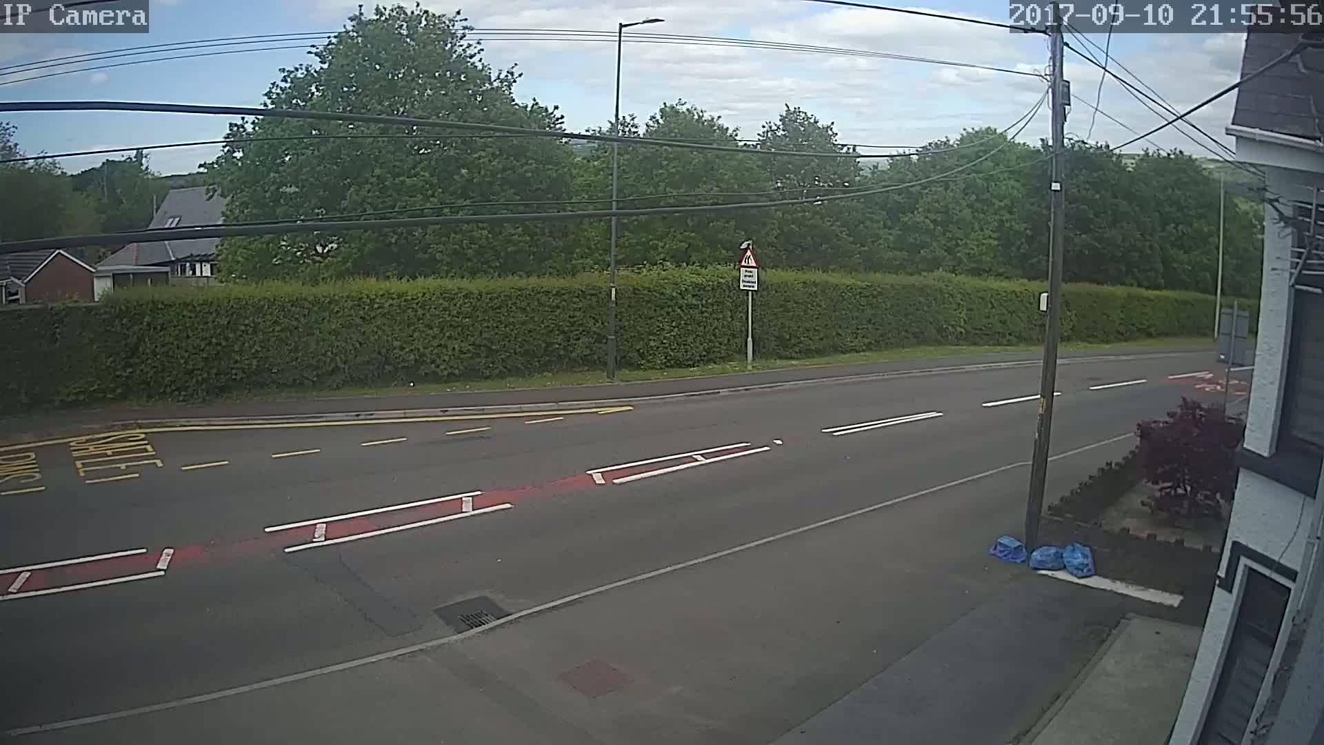 A mostly empty road with a pedestrian crossing and a tall hedge is visible under a partly cloudy sky.