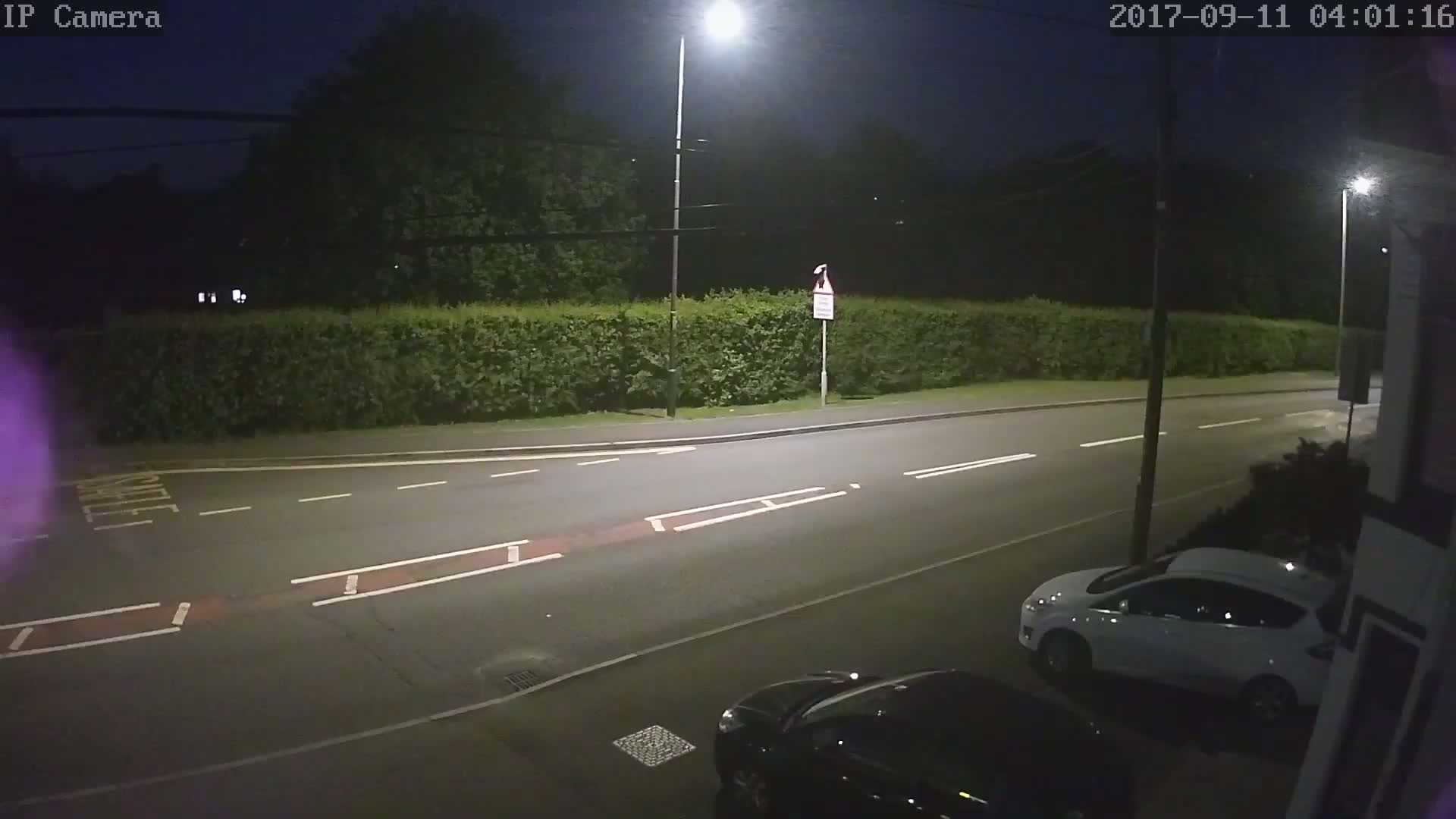 A nighttime view of a mostly empty road, lined with a hedge, illuminated by streetlights, with two cars parked near the side of the road.