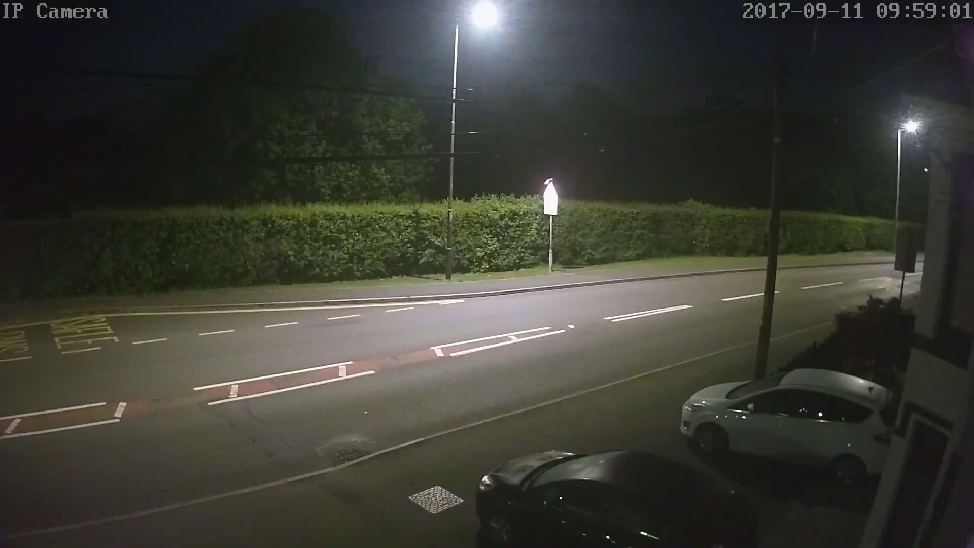 A nighttime view of a street with a white car parked and a dark-colored car partially visible, illuminated by streetlights, beside a hedge.
