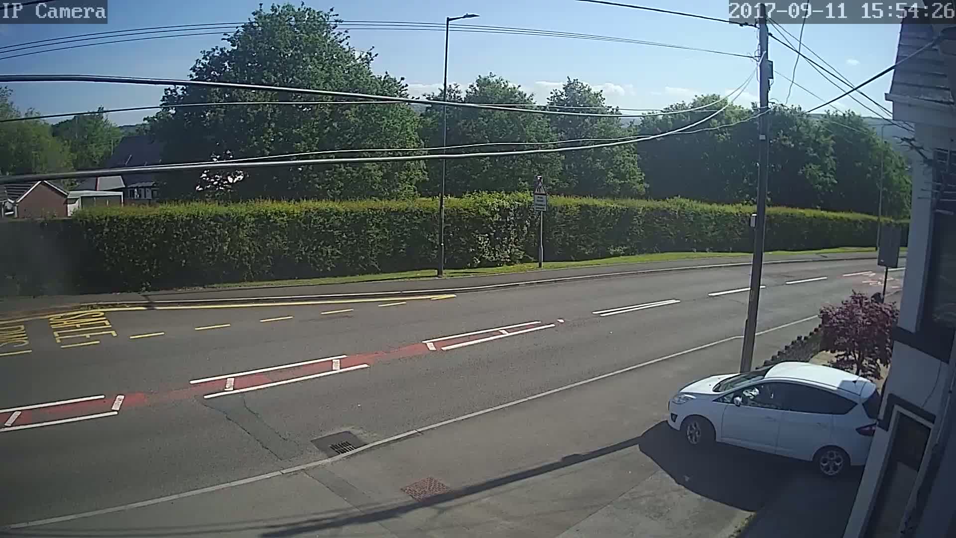 A white car is parked on the side of a road under a sunny sky, beside a tall hedge and a few trees.