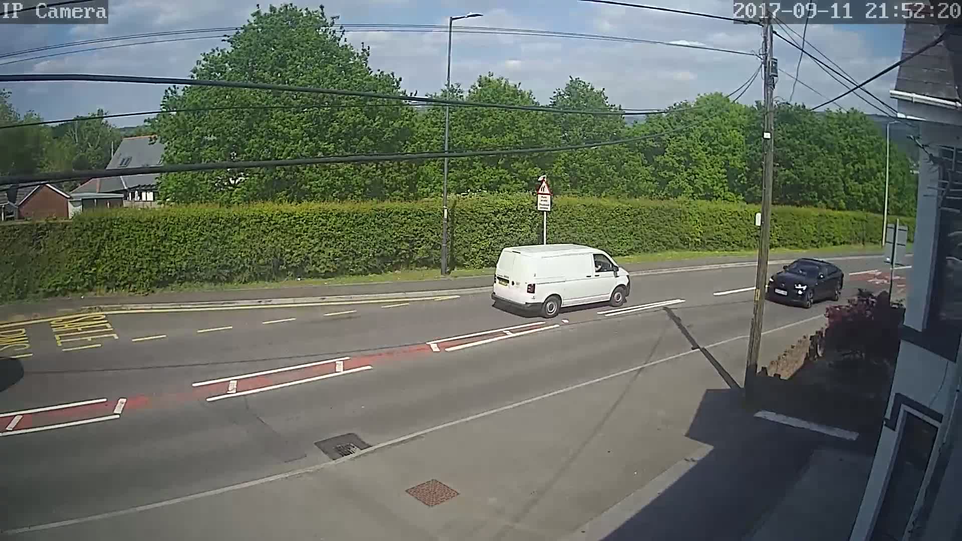 A white van and a dark-colored car are driving on a road under a partly sunny sky, past a green hedge and some trees.