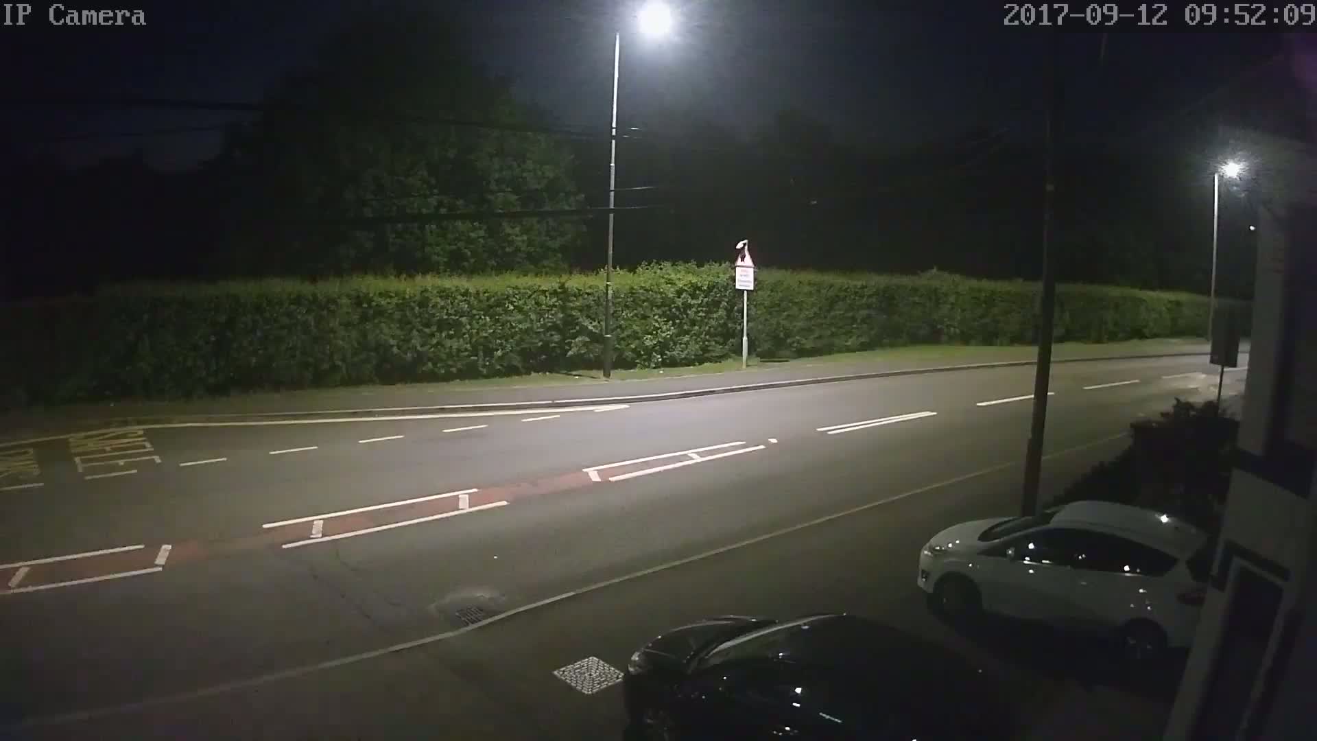 A nighttime view of a street illuminated by streetlights, showing a white car parked near a hedge and a dark-colored car partially visible in the foreground.