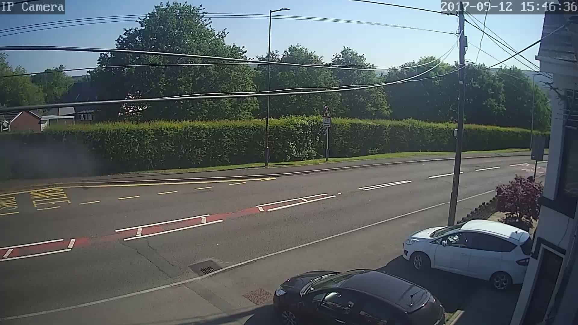 A sunny day reveals a street scene with two cars parked near a hedge, beyond which are trees and houses under a clear sky.